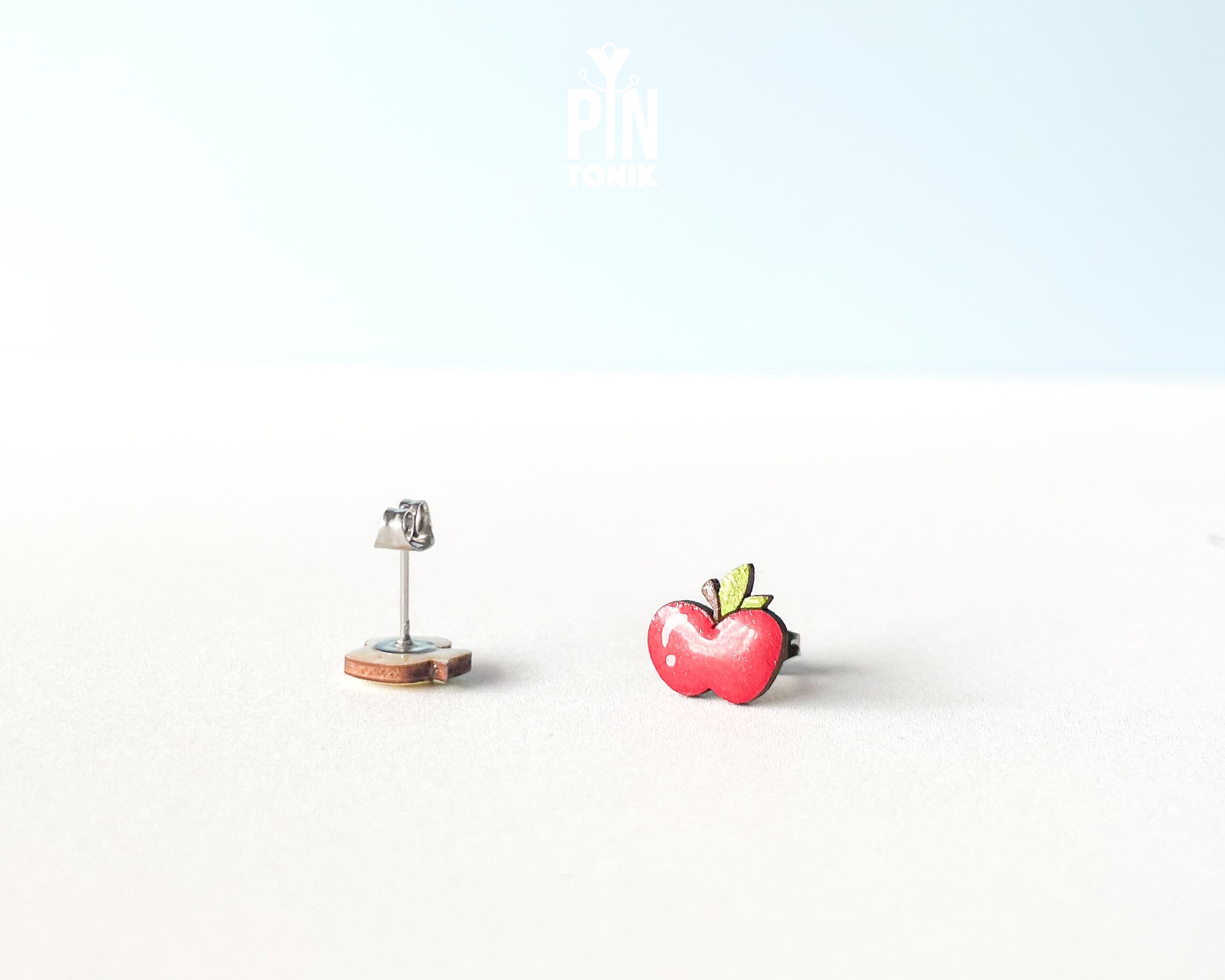Mismatched wooden stud earrings featuring one whole red apple and one yellow apple core illustration, high-gloss epoxy finish, shown on a white background.
