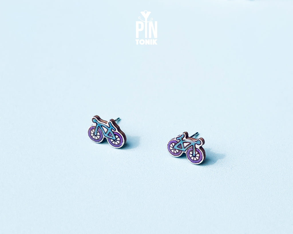 "Handmade wooden bicycle stud earrings illustrated in retro purple and blue shades by PinTonik."
