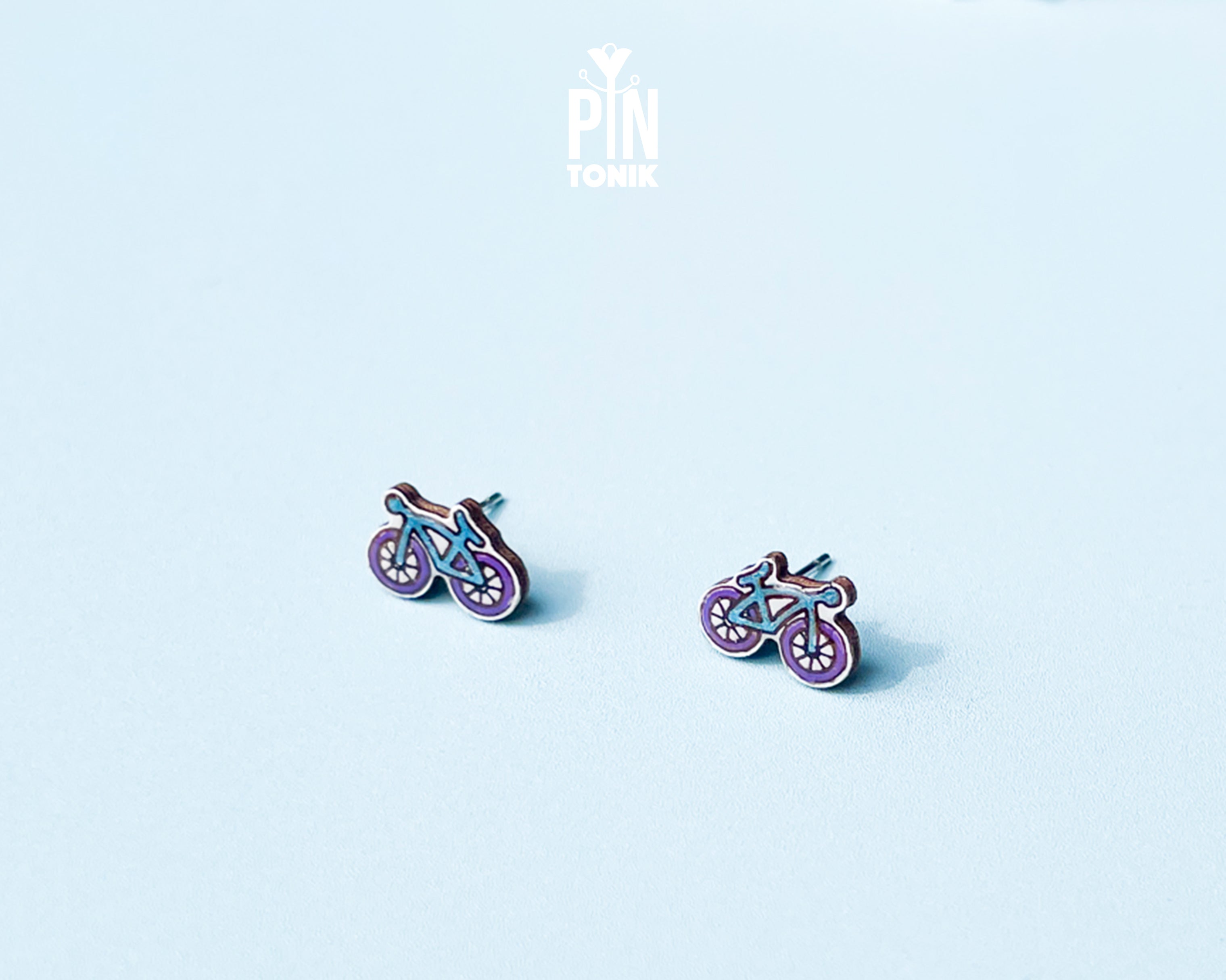 "Handmade wooden bicycle stud earrings illustrated in retro purple and blue shades by PinTonik."

