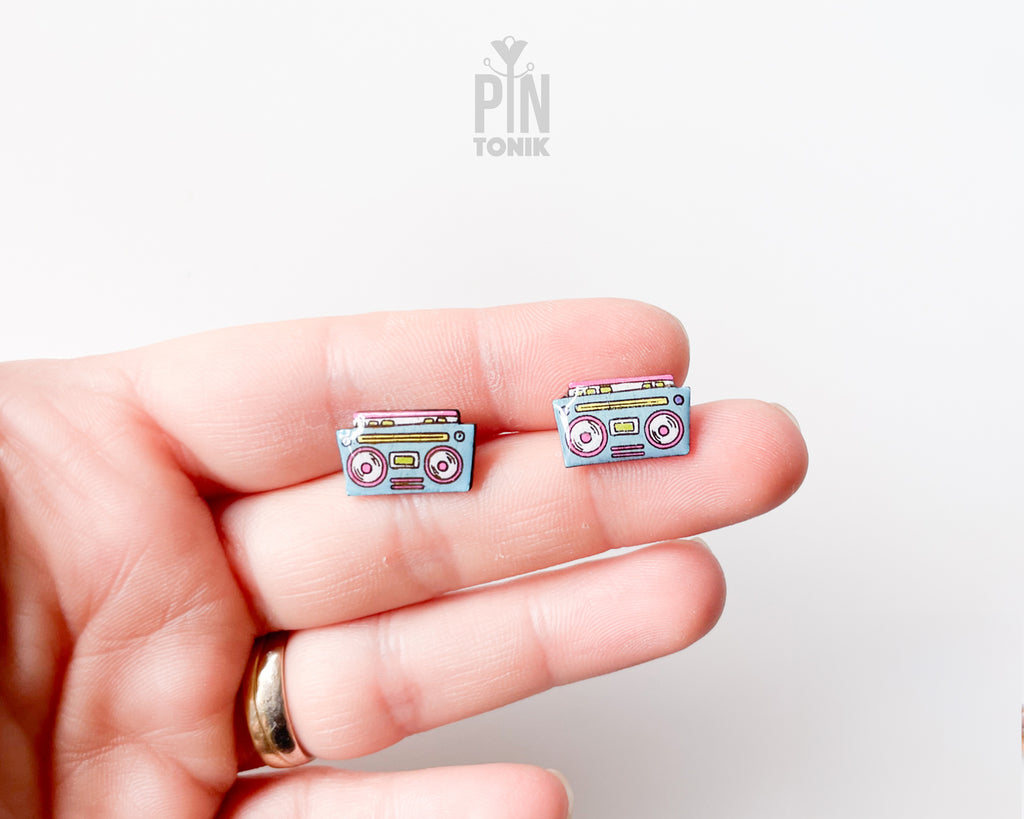 A close-up of a person's hand holding a pair of small cassette tape-shaped stud earrings. The earrings have a light blue-gray base with pink, yellow, and white details.