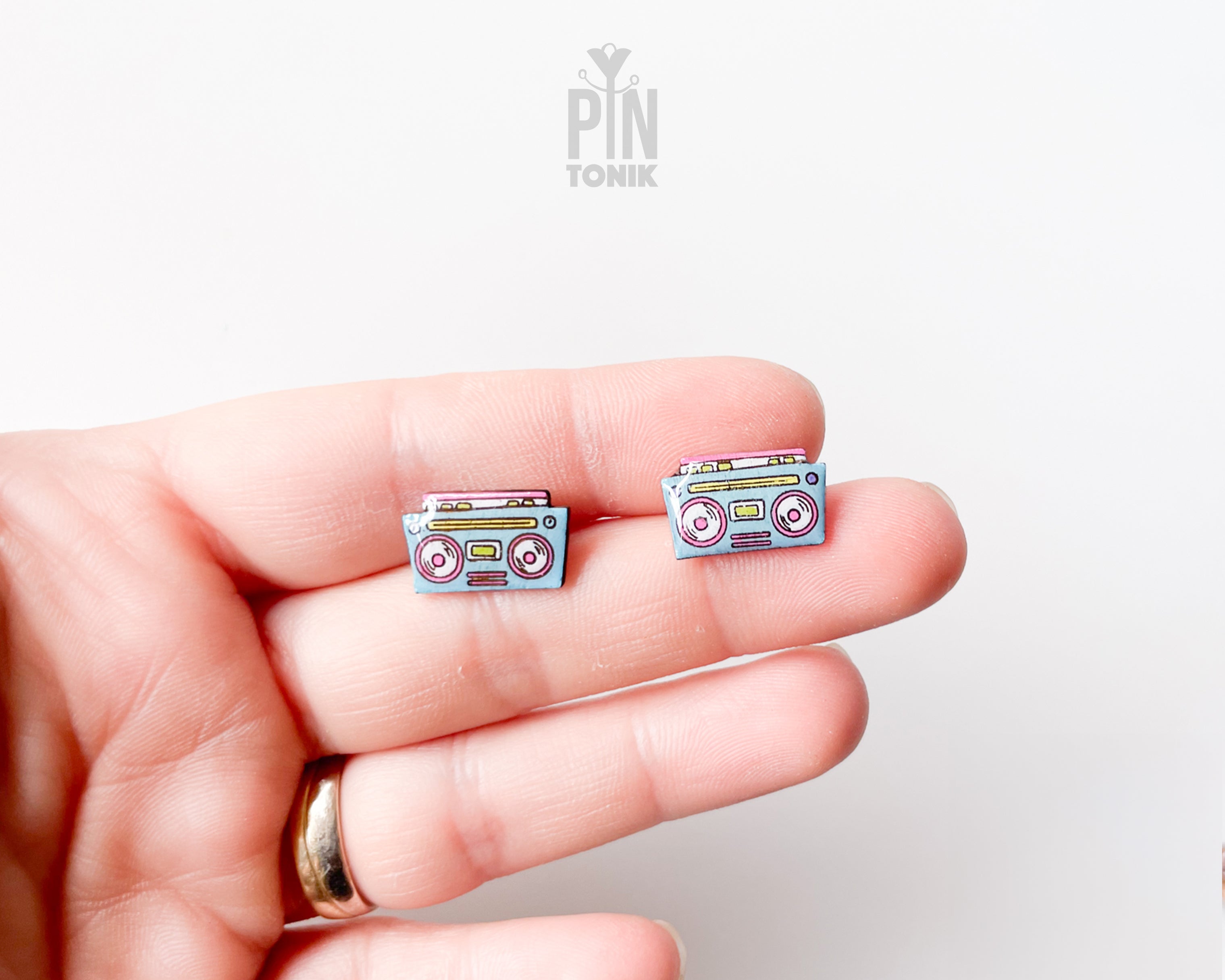 A close-up of a person's hand holding a pair of small cassette tape-shaped stud earrings. The earrings have a light blue-gray base with pink, yellow, and white details.