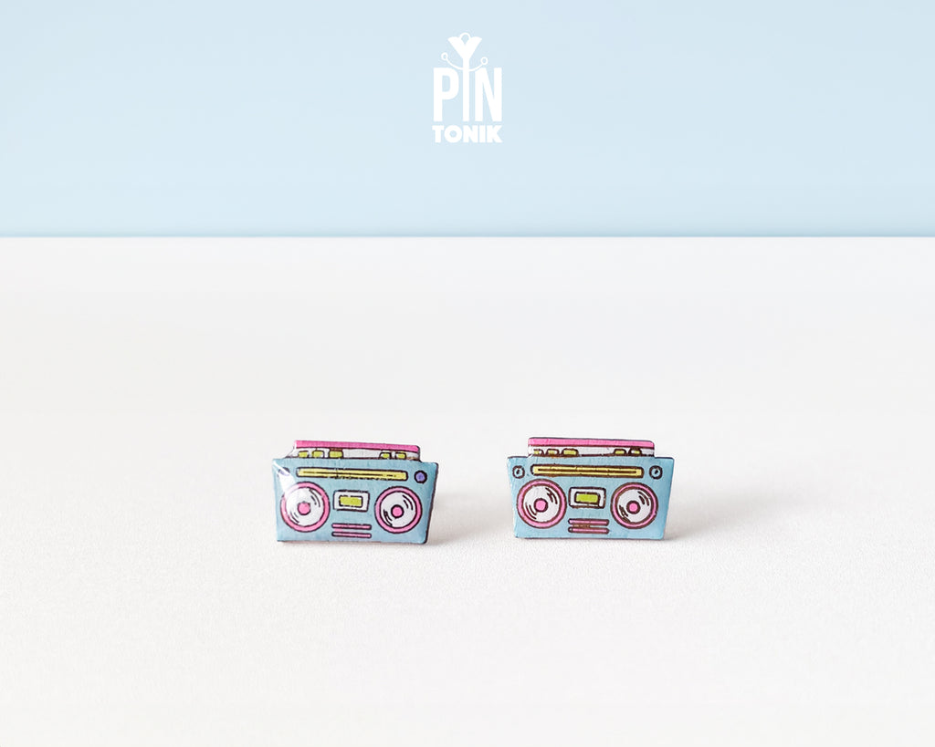A pair of retro cassette tape stud earrings on a white surface with a light blue background. The earrings are light blue with pink, yellow, and red details representing the cassette tape design.