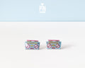 A pair of retro cassette tape stud earrings on a white surface with a light blue background. The earrings are light blue with pink, yellow, and red details representing the cassette tape design.