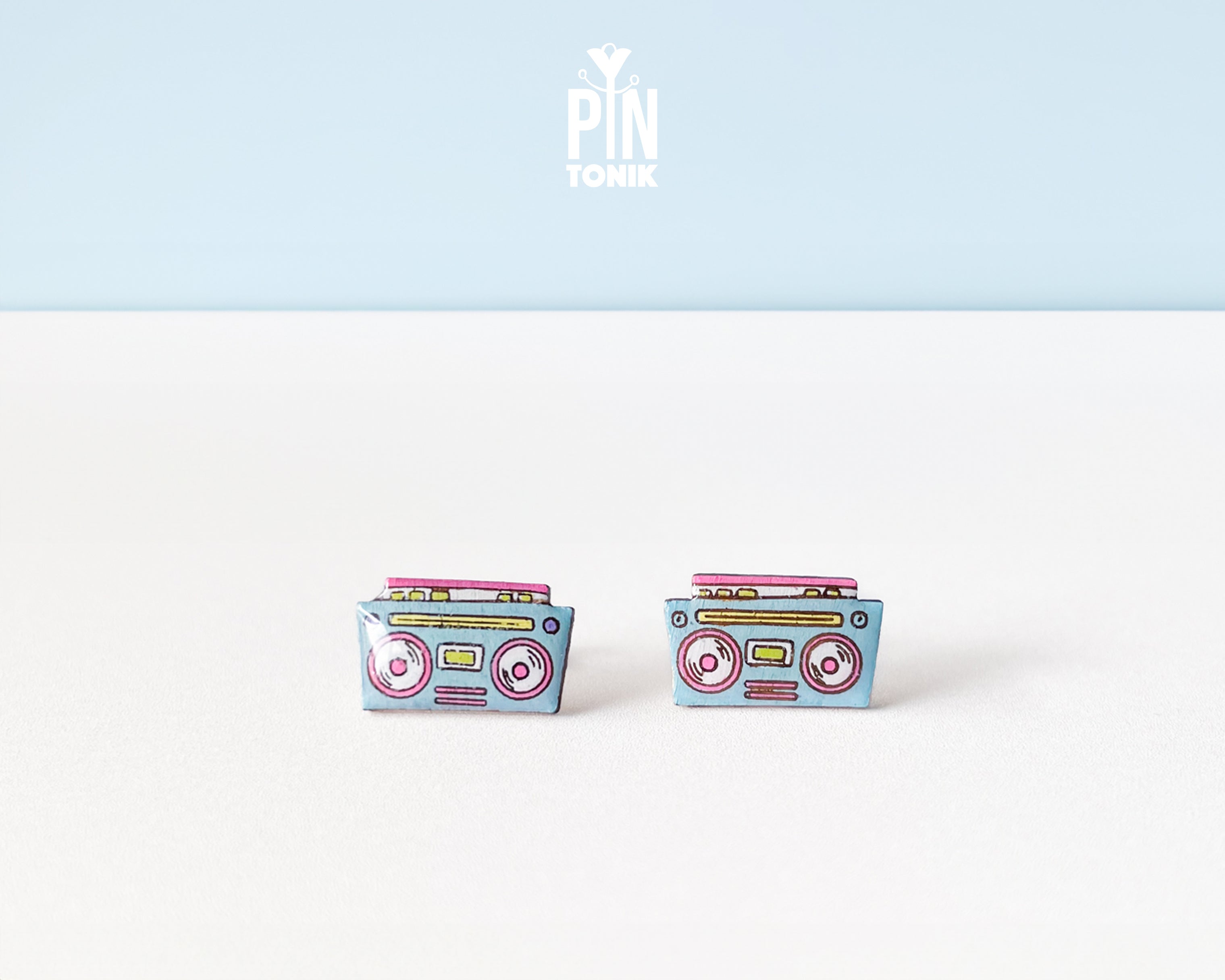 A pair of retro cassette tape stud earrings on a white surface with a light blue background. The earrings are light blue with pink, yellow, and red details representing the cassette tape design.