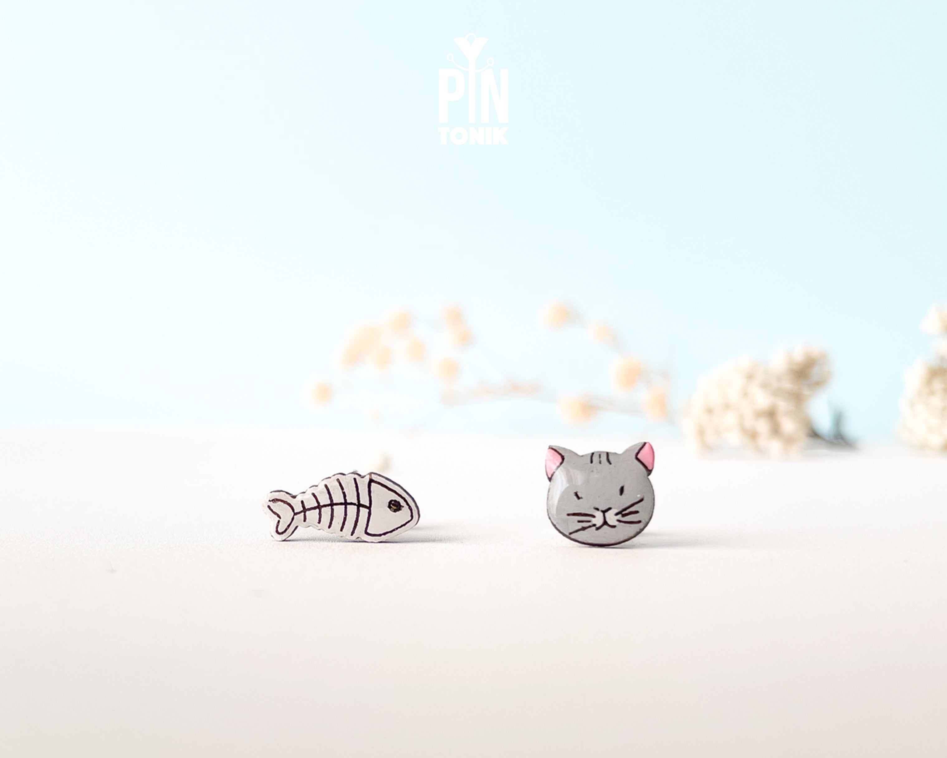 Mismatched hand-painted wooden stud earrings featuring a grey cat face and a white fishbone skeleton