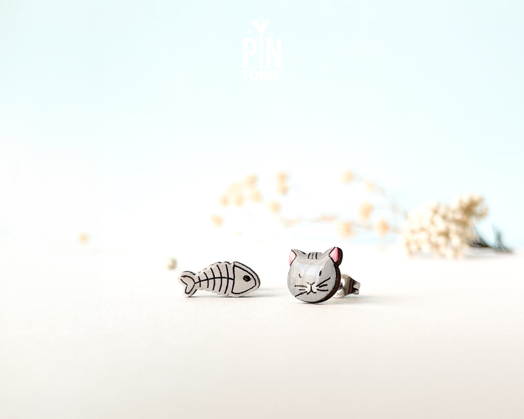 Mismatched hand-painted wooden stud earrings featuring a grey cat face and a white fishbone skeleton
