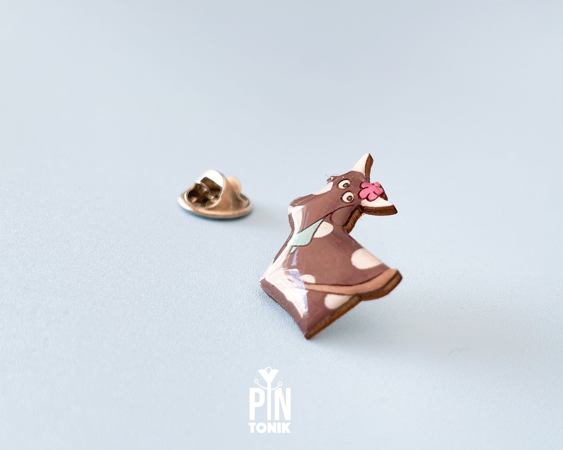 Hand-painted brown and white wooden cow pin with a pink flower and a glossy resin finish