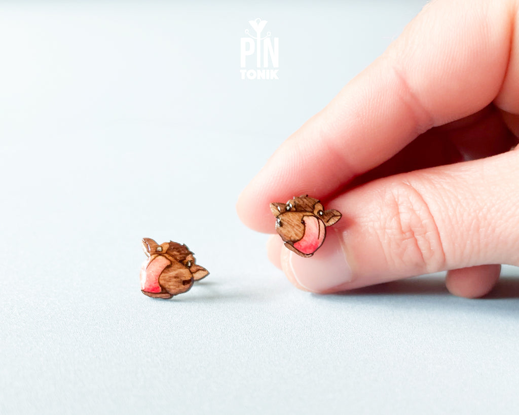 A close-up of a hand holding a small animal-shaped stud earring next to another matching earring on a light gray background. The earrings feature a brown wood-grain texture with pink enamel accents.