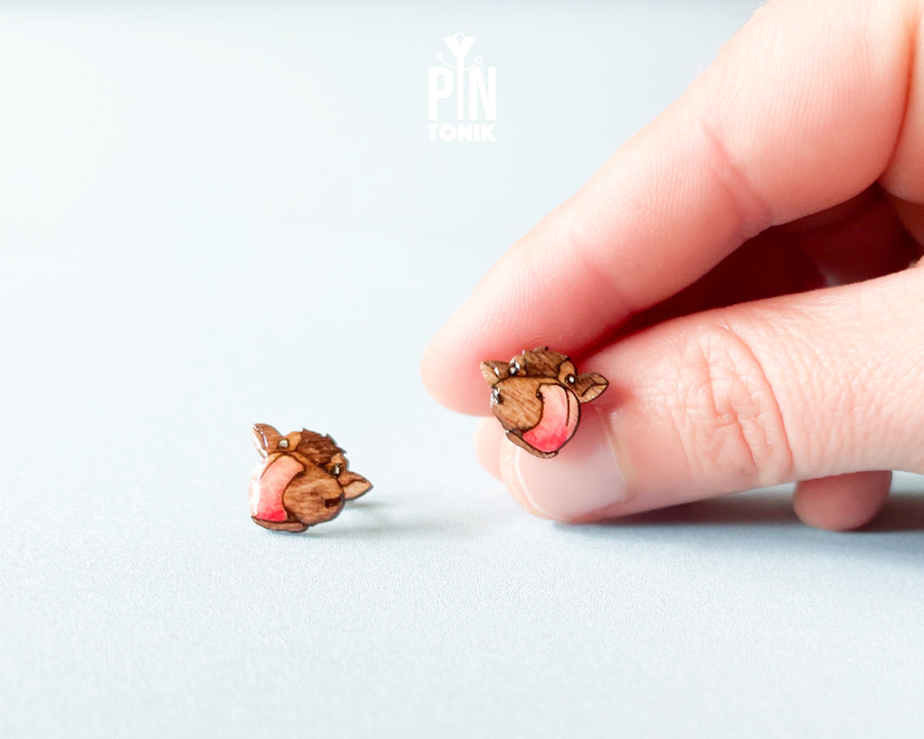A close-up of a hand holding a small animal-shaped stud earring next to another matching earring on a light gray background. The earrings feature a brown wood-grain texture with pink enamel accents.