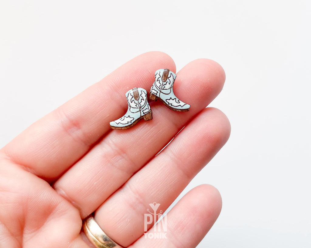 A close-up of a hand holding a pair of small cowboy boot stud earrings. The earrings are light blue with white and light brown details, shown against a plain white background.