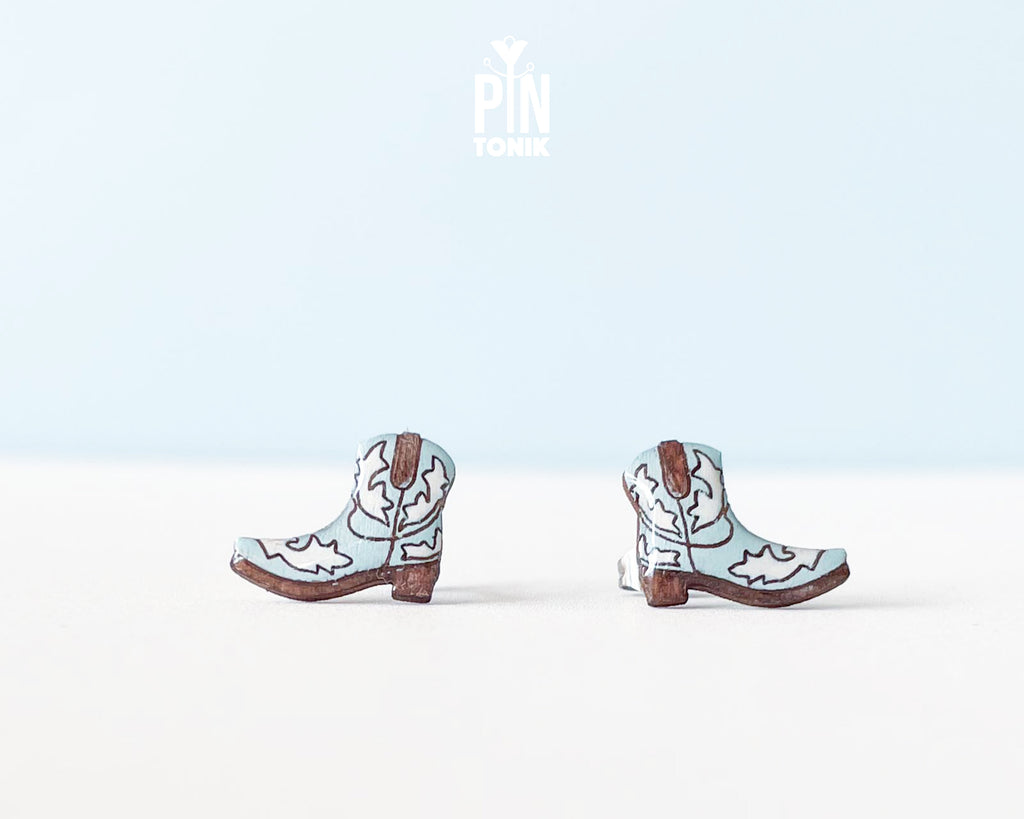 A pair of miniature cowboy boot earrings on a clean white surface with a light blue background. The boots are light blue with brown heels, soles, and stitching details, and have a stainless steel post visible on the back.