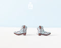 A pair of miniature cowboy boot earrings on a clean white surface with a light blue background. The boots are light blue with brown heels, soles, and stitching details, and have a stainless steel post visible on the back.
