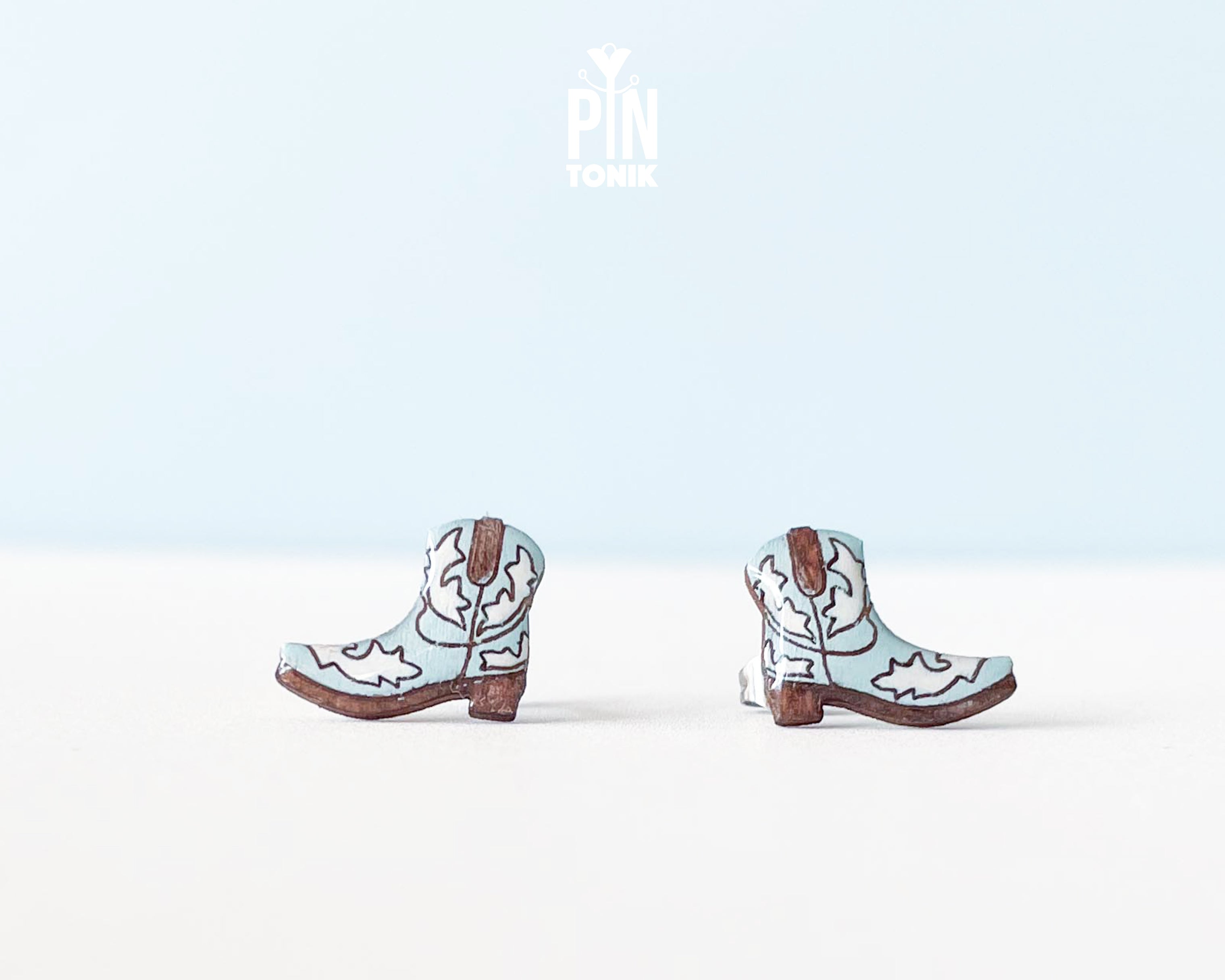A pair of miniature cowboy boot earrings on a clean white surface with a light blue background. The boots are light blue with brown heels, soles, and stitching details, and have a stainless steel post visible on the back.