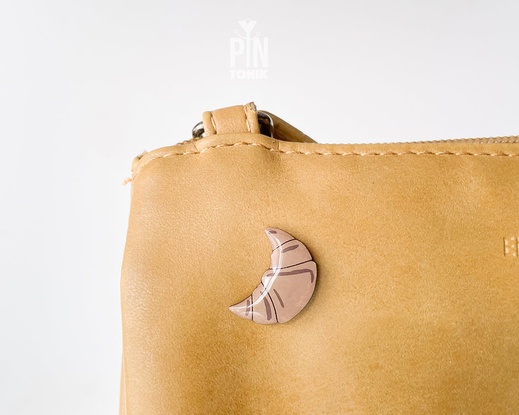 Hand-painted wooden croissant pin with golden-brown layers and a high-gloss finish, shown next to a stainless steel clutch back on a light blue background.