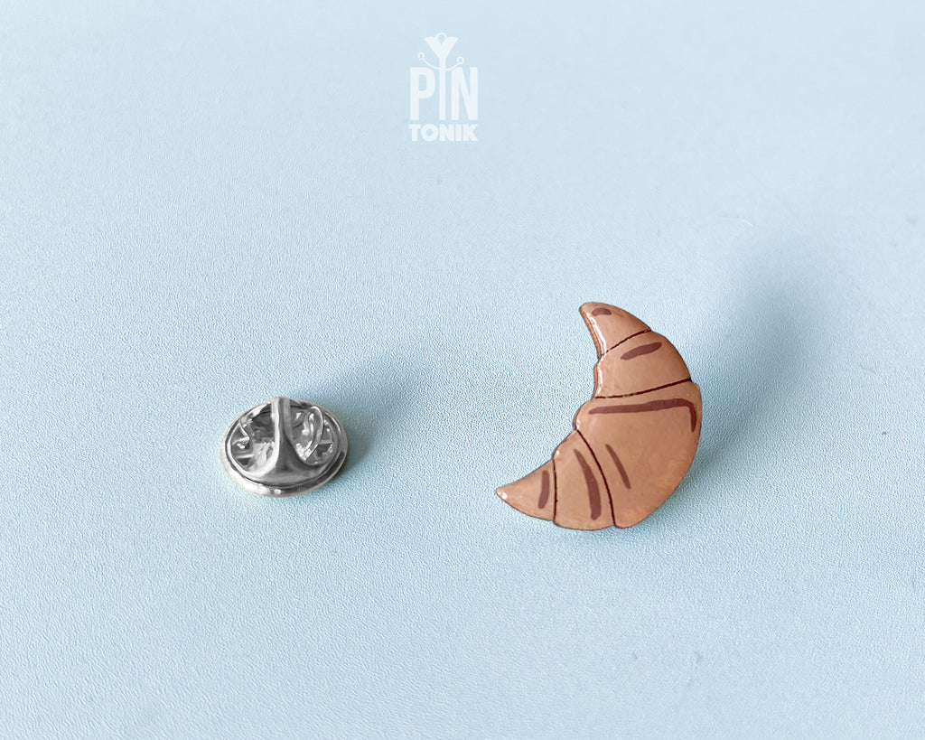 Hand-painted wooden croissant pin with golden-brown layers and a high-gloss finish, shown next to a stainless steel clutch back on a light blue background.
