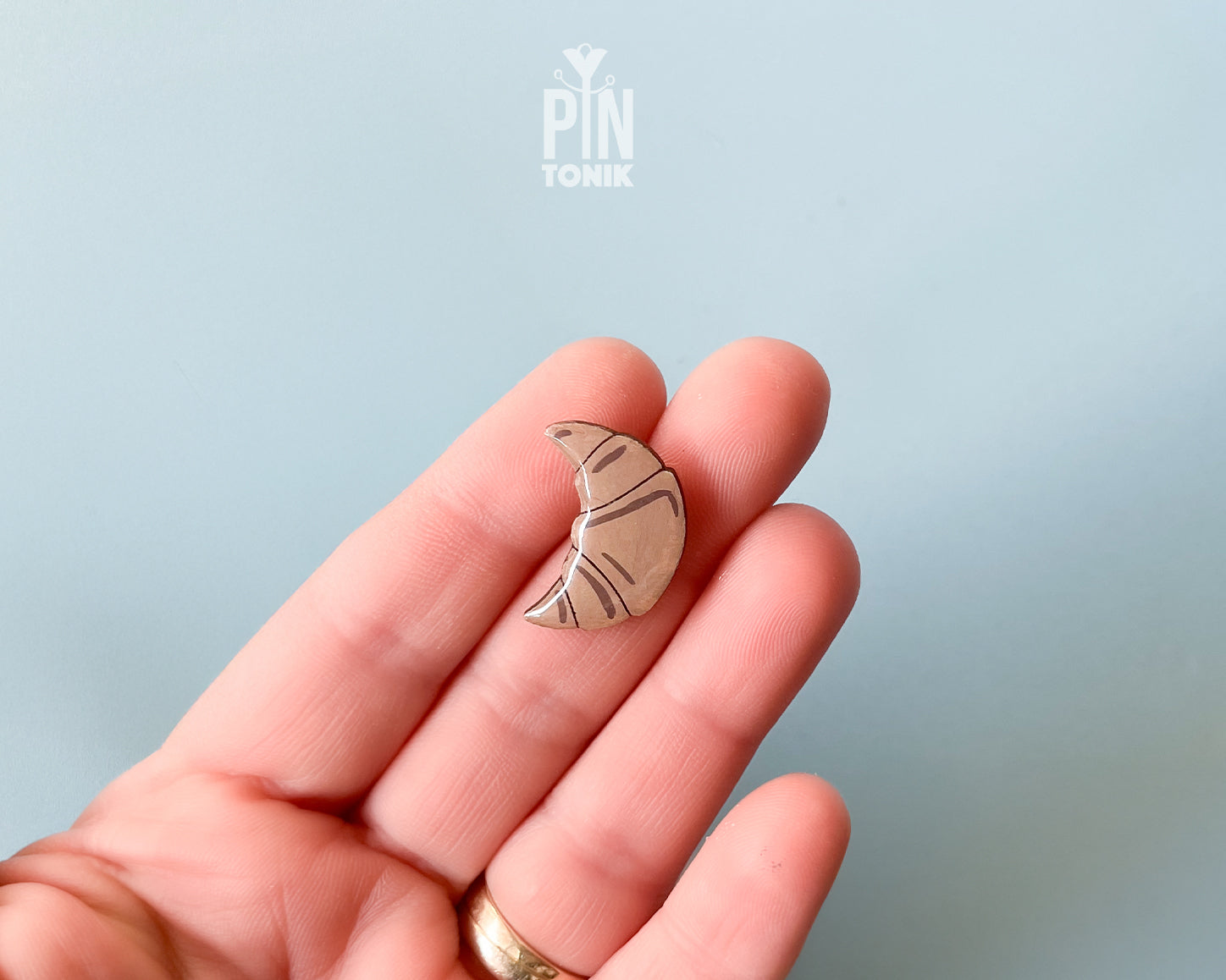 Hand-painted wooden croissant pin with golden-brown layers and a high-gloss finish, shown next to a stainless steel clutch back on a light blue background.