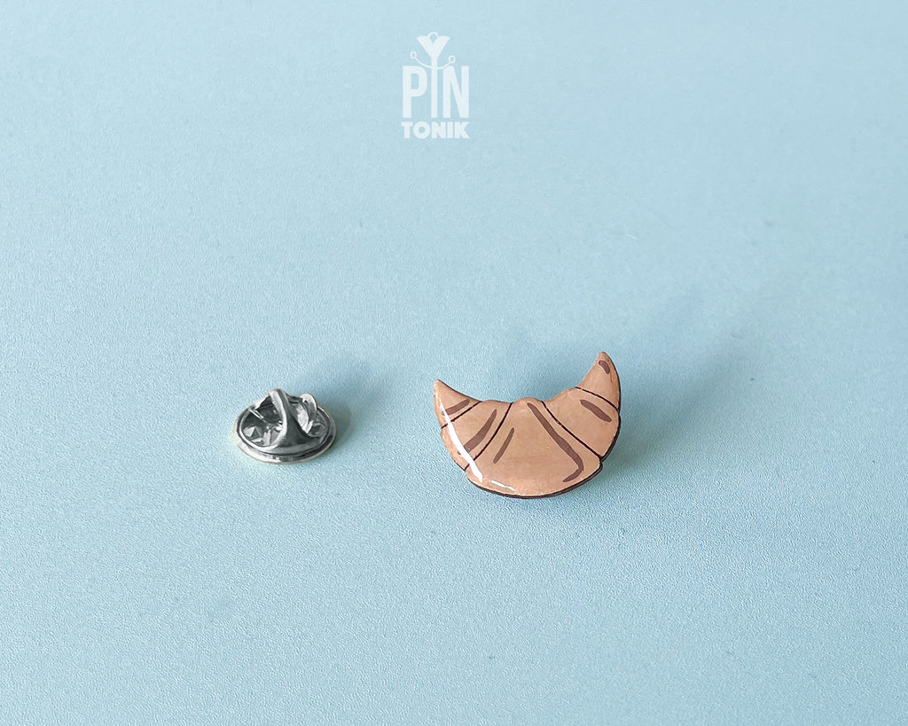 Hand-painted wooden croissant pin with golden-brown layers and a high-gloss finish, shown next to a stainless steel clutch back on a light blue background.