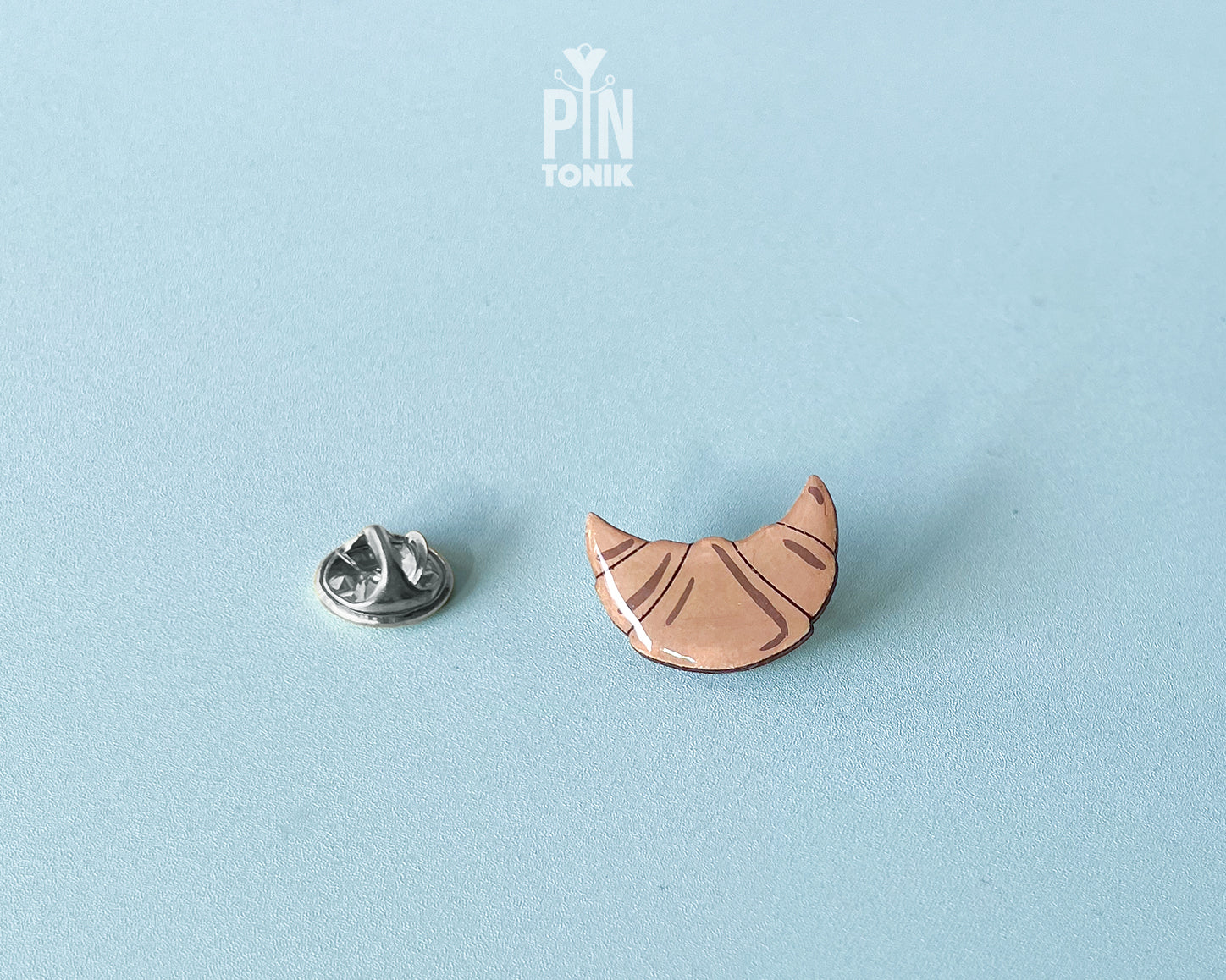 Hand-painted wooden croissant pin with golden-brown layers and a high-gloss finish, shown next to a stainless steel clutch back on a light blue background.