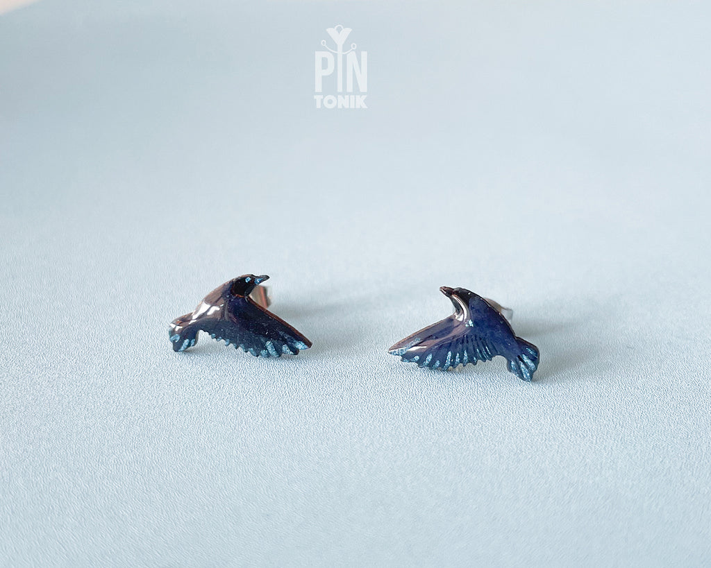 A pair of small raven stud earrings with dark blue and black enamel and subtle metallic highlights, displayed on a light blue textured surface. A faint 'PIN TONIK' watermark is visible at the top center.