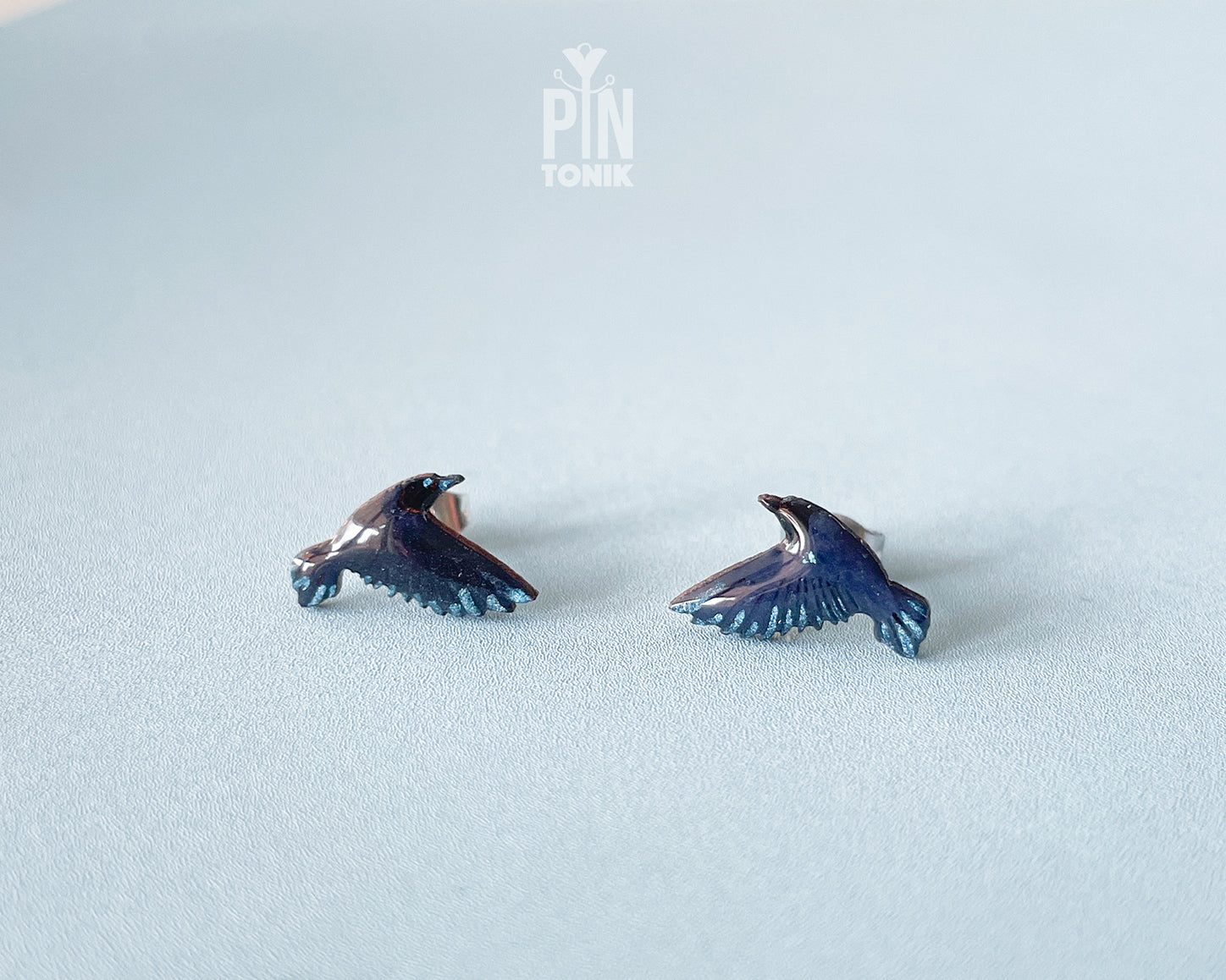 A pair of small raven stud earrings with dark blue and black enamel and subtle metallic highlights, displayed on a light blue textured surface. A faint 'PIN TONIK' watermark is visible at the top center.