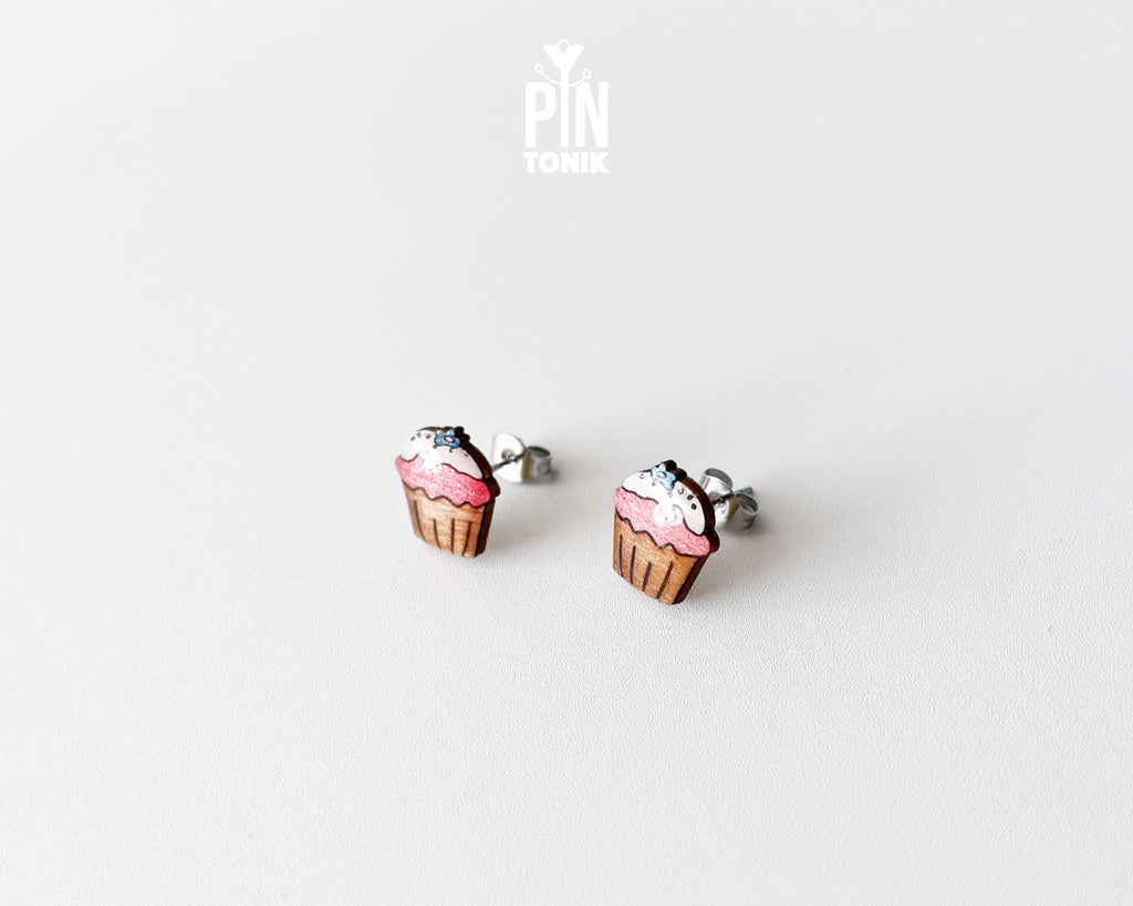 A pair of stud earrings shaped like cupcakes with pink frosting, brown liners, and white sprinkles, shown on a plain light gray background. A small 'PIN TONIK' logo is visible near the top center.