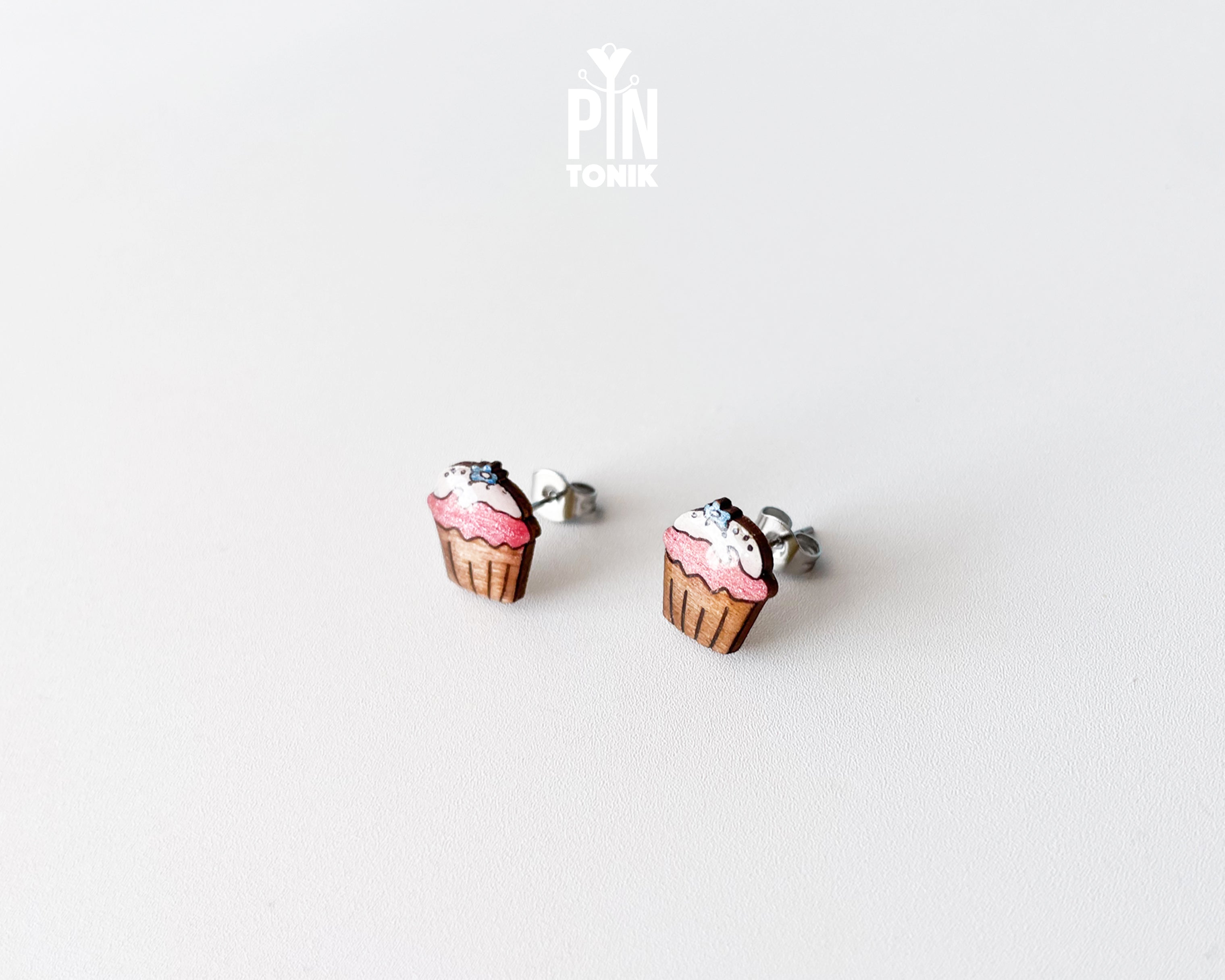 A pair of stud earrings shaped like cupcakes with pink frosting, brown liners, and white sprinkles, shown on a plain light gray background. A small 'PIN TONIK' logo is visible near the top center.