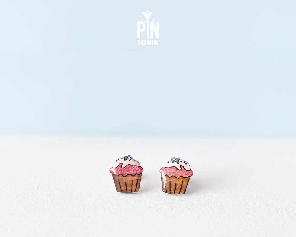 A pair of cupcake-shaped stud earrings with pink frosting, white icing, and blue sprinkles, displayed on a clean white surface against a light blue background. The brand logo 'PIN TONIK' is visible at the top center.