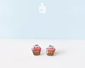 A pair of cupcake-shaped stud earrings with pink frosting, white icing, and blue sprinkles, displayed on a clean white surface against a light blue background. The brand logo 'PIN TONIK' is visible at the top center.