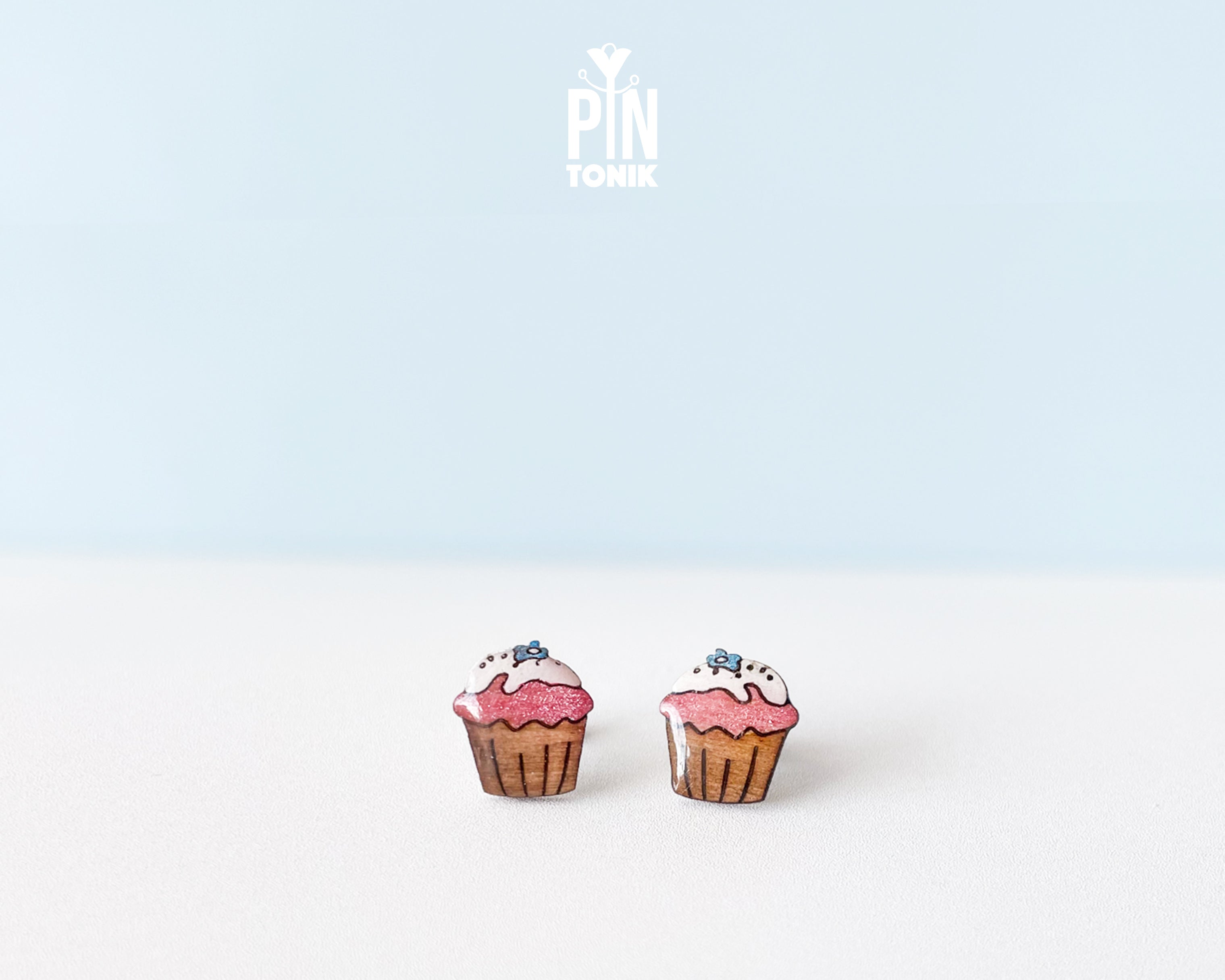 A pair of cupcake-shaped stud earrings with pink frosting, white icing, and blue sprinkles, displayed on a clean white surface against a light blue background. The brand logo 'PIN TONIK' is visible at the top center.