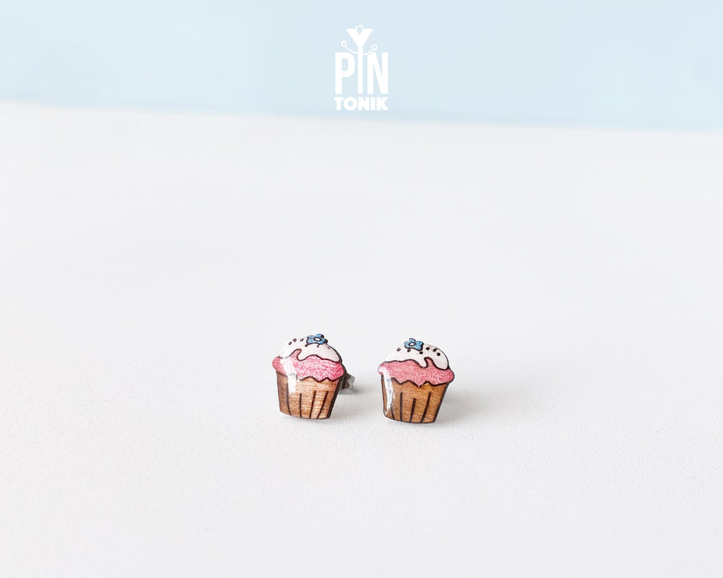 A pair of cupcake stud earrings on a clean white surface with a light blue background. The earrings depict pink frosted cupcakes with brown liners and small blue sprinkles.