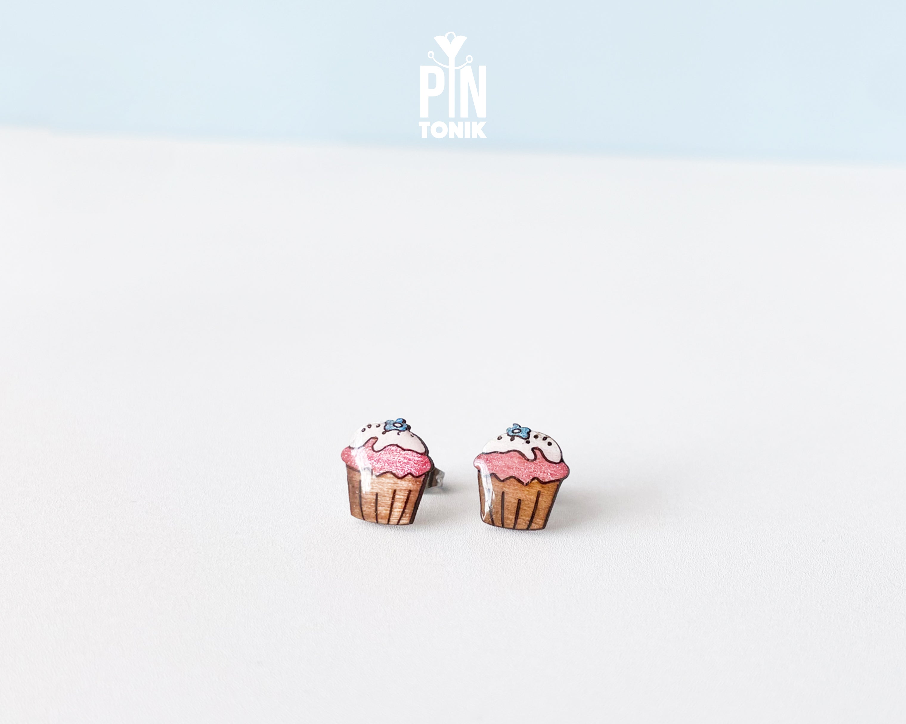 A pair of cupcake stud earrings on a clean white surface with a light blue background. The earrings depict pink frosted cupcakes with brown liners and small blue sprinkles.