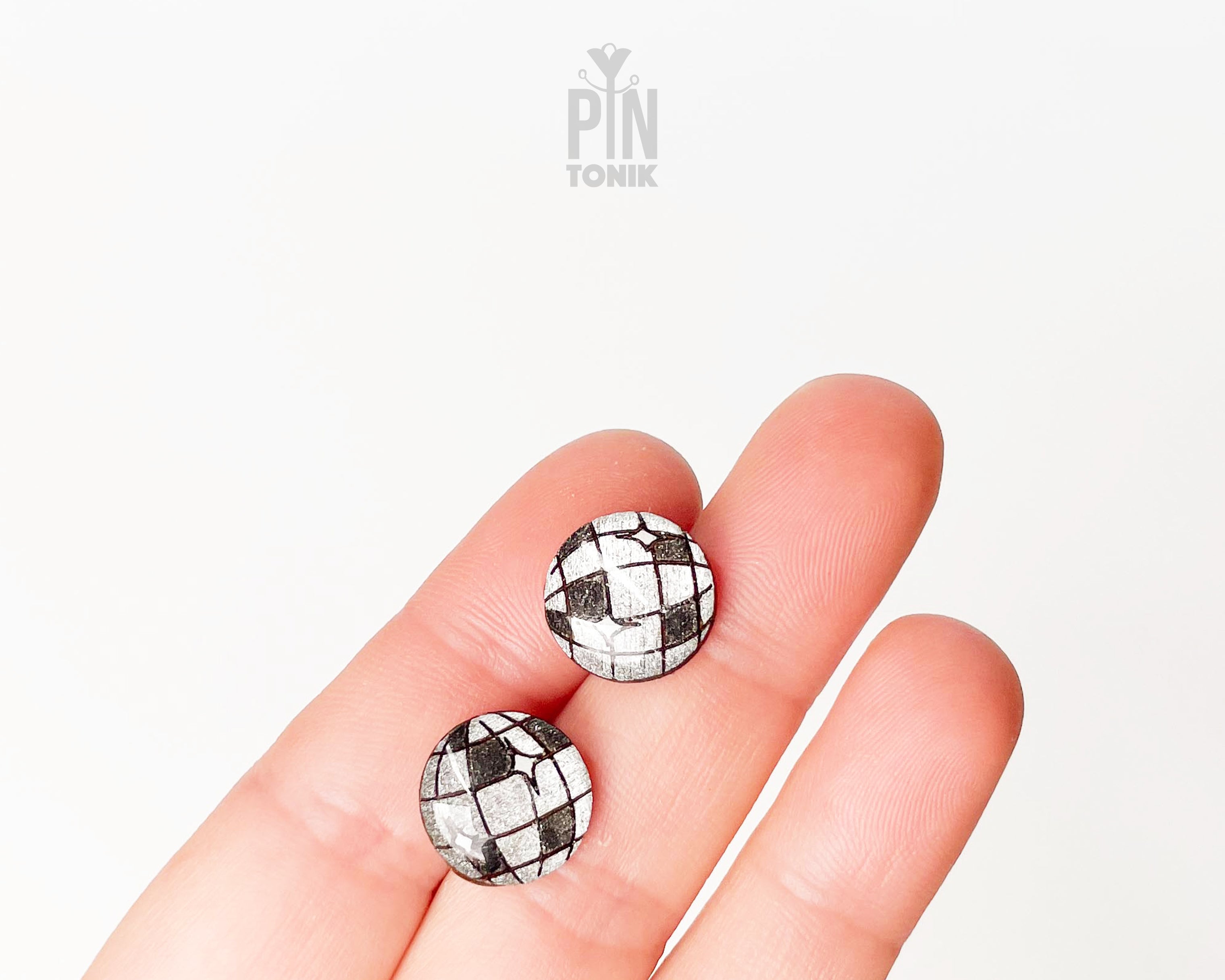 A close-up of a person's fingers holding two small round wooden stud earrings with a Silver and Anthracite  checkered disco ball pattern against a plain white background.