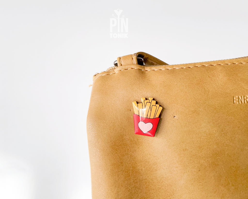 Hand-painted wooden french fries pin in a red carton with a small pink heart, shown next to a stainless steel clutch back on a light blue background.
