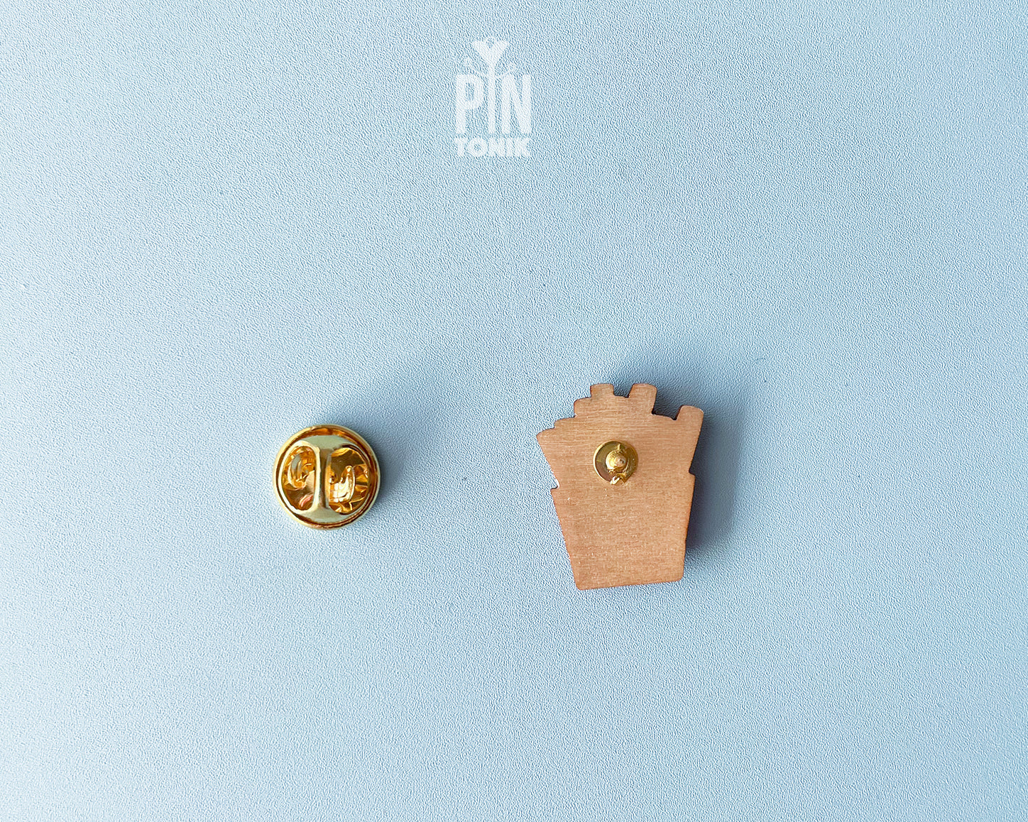 Hand-painted wooden french fries pin in a red carton with a small pink heart, shown next to a stainless steel clutch back on a light blue background.