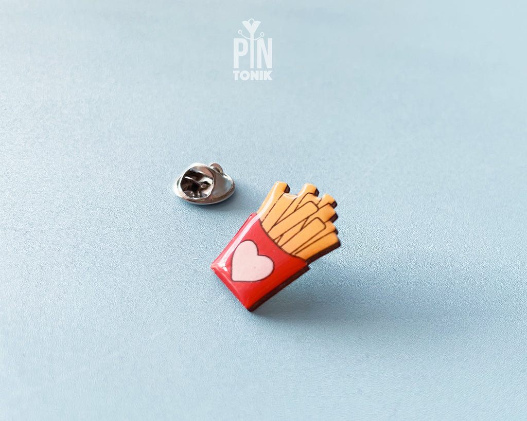 Hand-painted wooden french fries pin in a red carton with a small pink heart, shown next to a stainless steel clutch back on a light blue background.