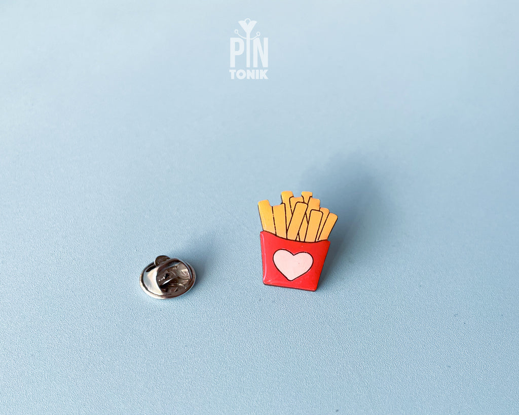 Hand-painted wooden french fries pin in a red carton with a small pink heart, shown next to a stainless steel clutch back on a light blue background.