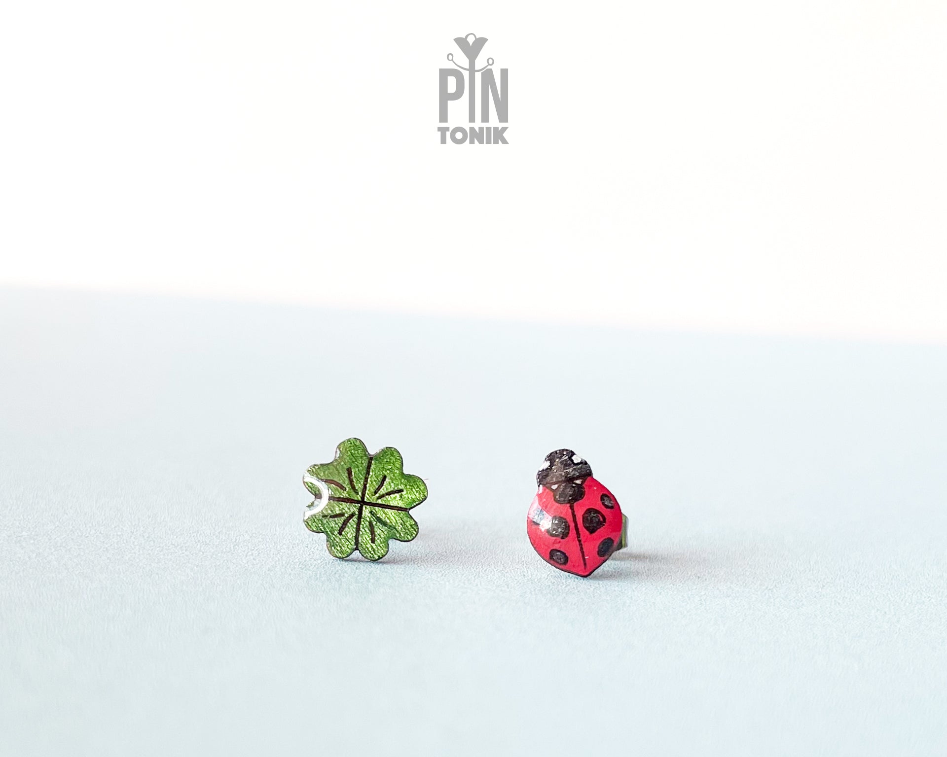 Handmade wooden stud earrings featuring a tiny red ladybug sitting on a green four-leaf clover by PinTonik.
