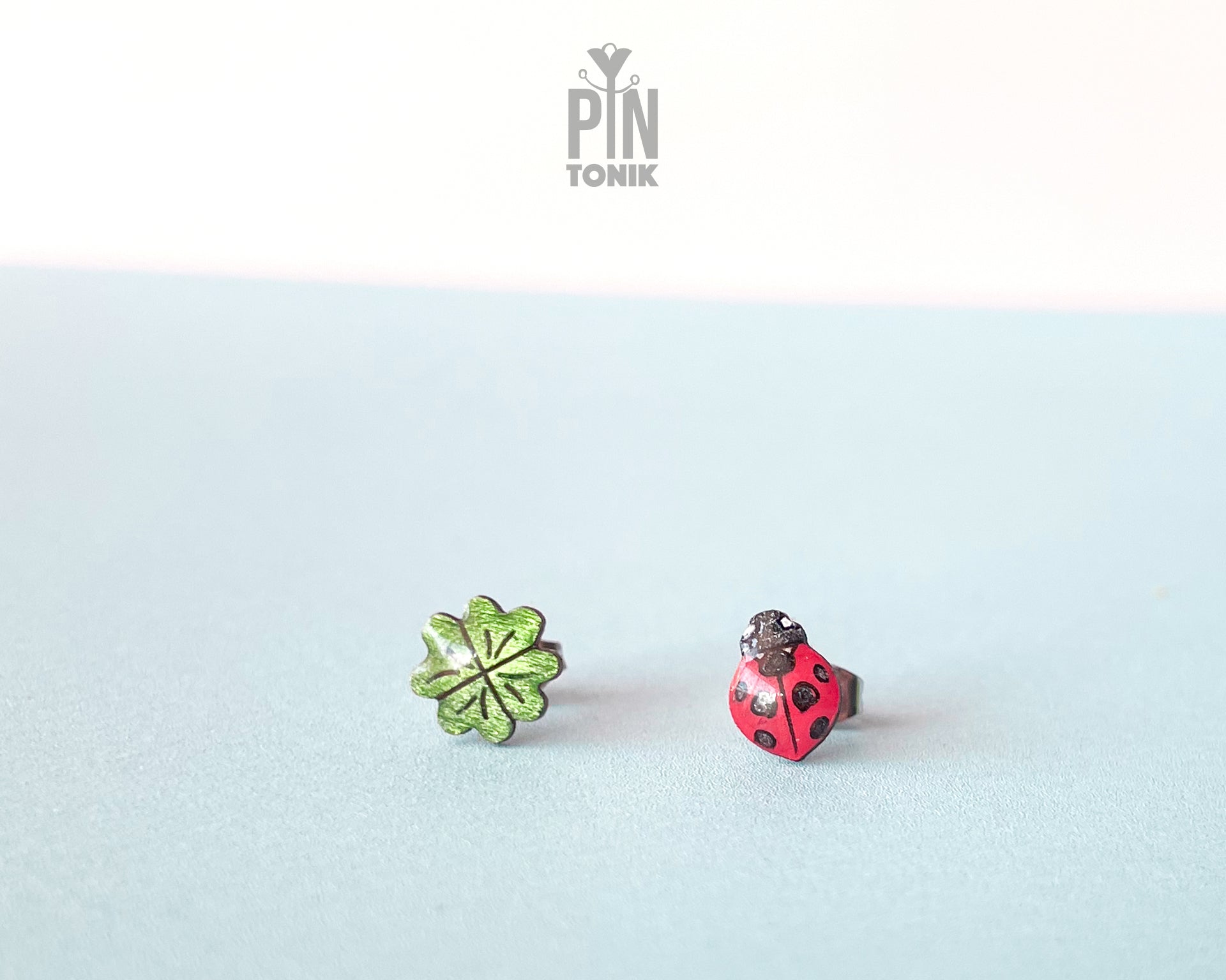 Handmade wooden stud earrings featuring a tiny red ladybug sitting on a green four-leaf clover by PinTonik.