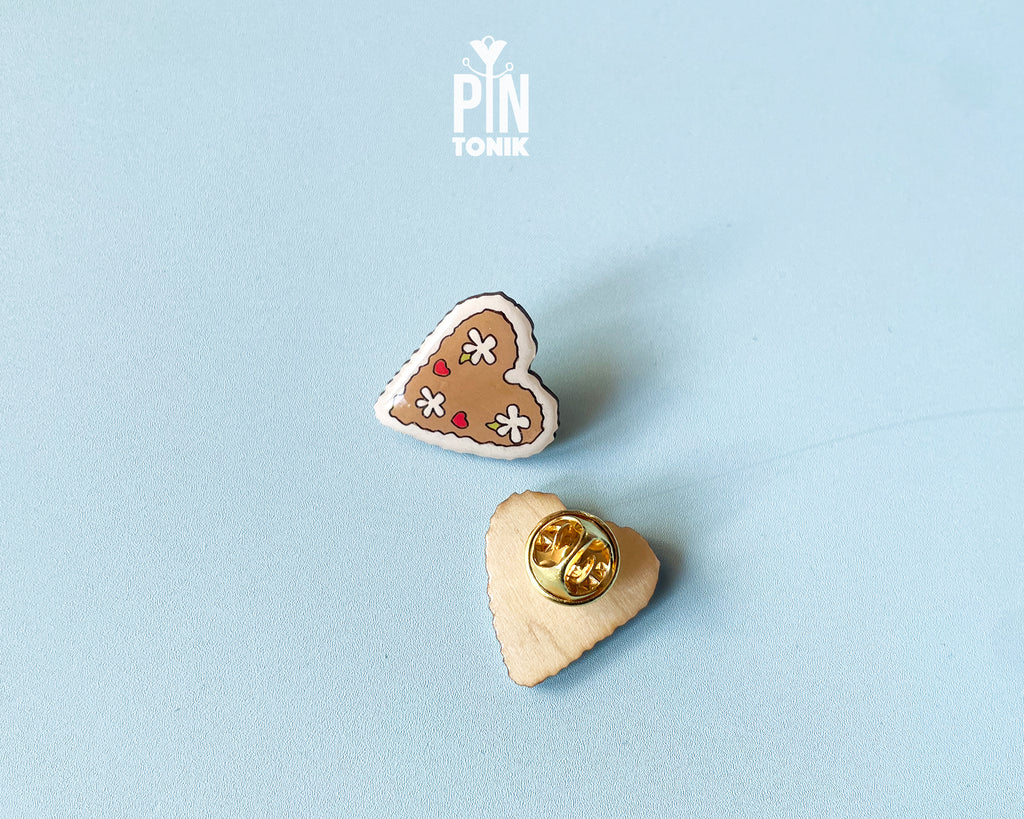 Hand-painted wooden gingerbread heart pins with white "icing" edges and floral details. One version features the Tyrolean text "I mag Di" while the other is a simple floral heart.
