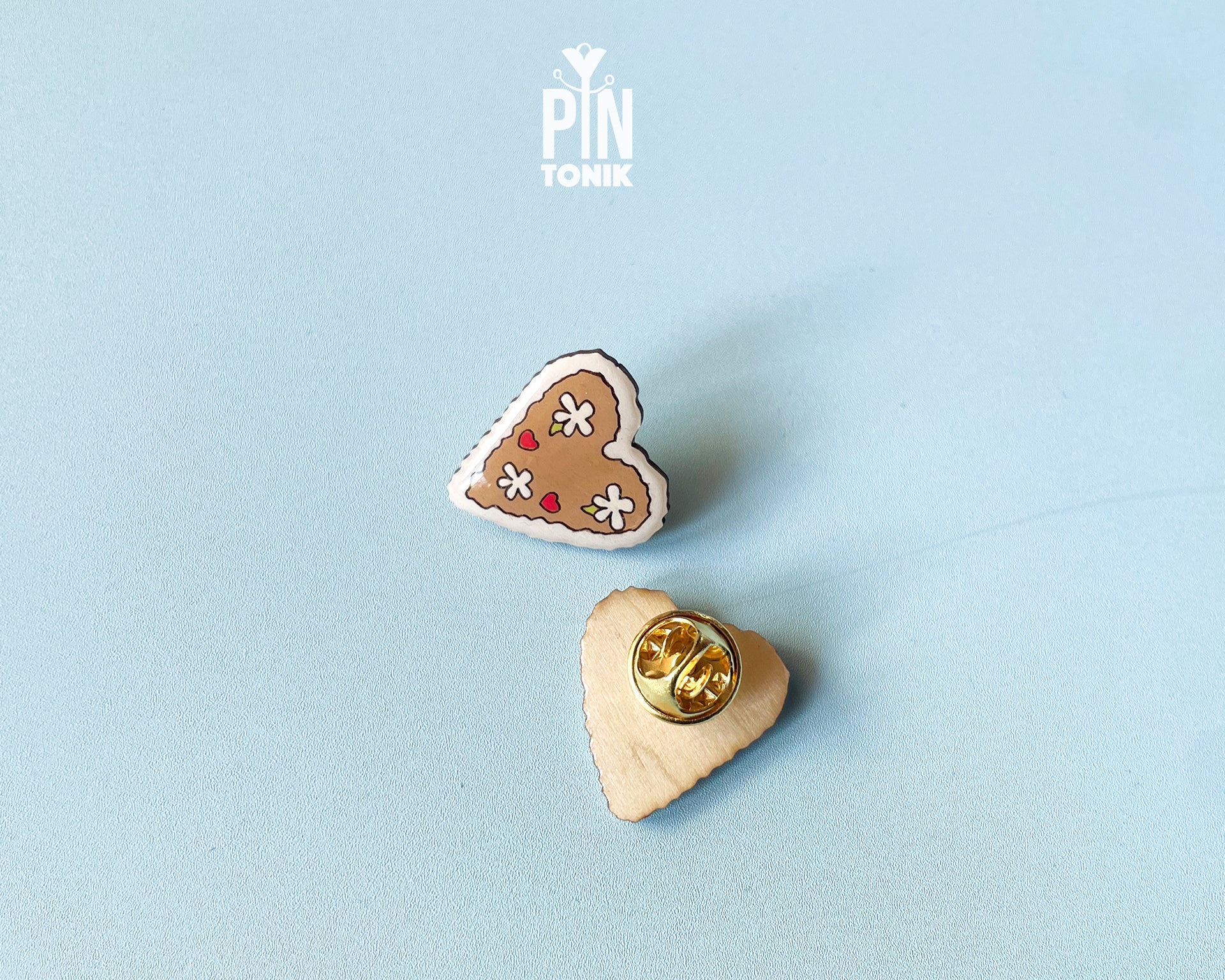 Hand-painted wooden gingerbread heart pins with white "icing" edges and floral details. One version features the Tyrolean text "I mag Di" while the other is a simple floral heart.