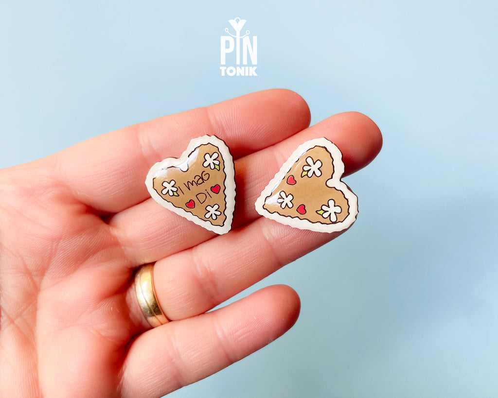 Hand-painted wooden gingerbread heart pins with white "icing" edges and floral details. One version features the Tyrolean text "I mag Di" while the other is a simple floral heart.