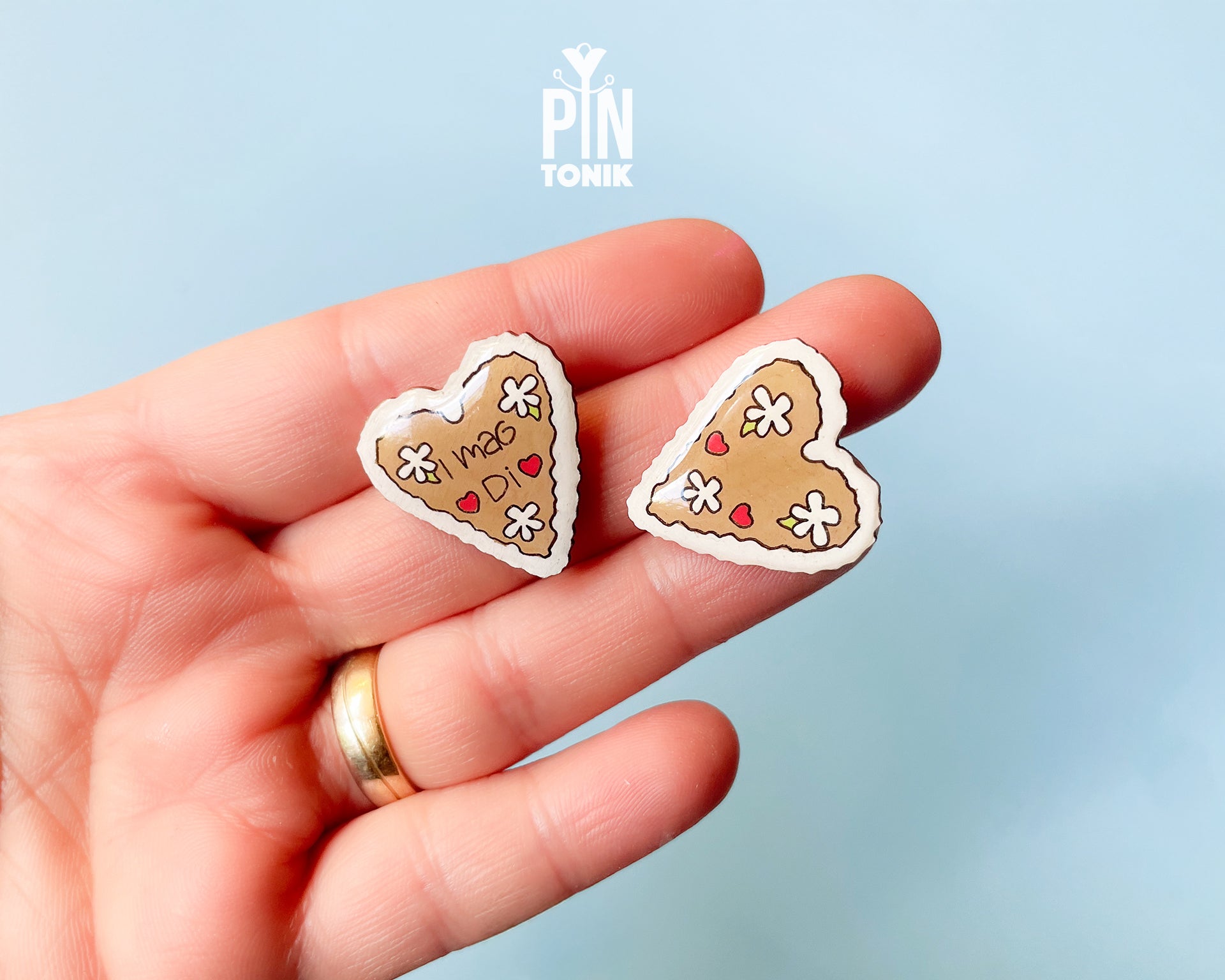 Hand-painted wooden gingerbread heart pins with white "icing" edges and floral details. One version features the Tyrolean text "I mag Di" while the other is a simple floral heart.