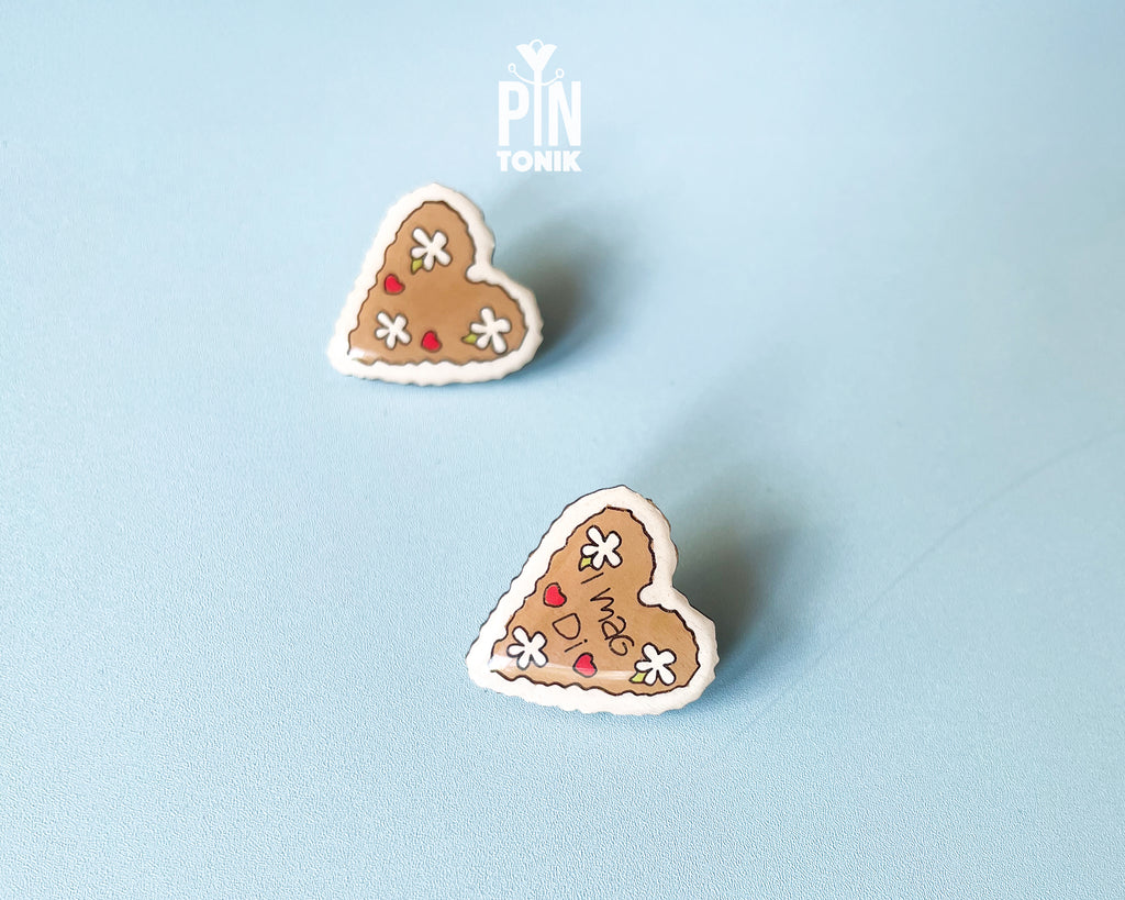 Hand-painted wooden gingerbread heart pins with white "icing" edges and floral details. One version features the Tyrolean text "I mag Di" while the other is a simple floral heart.