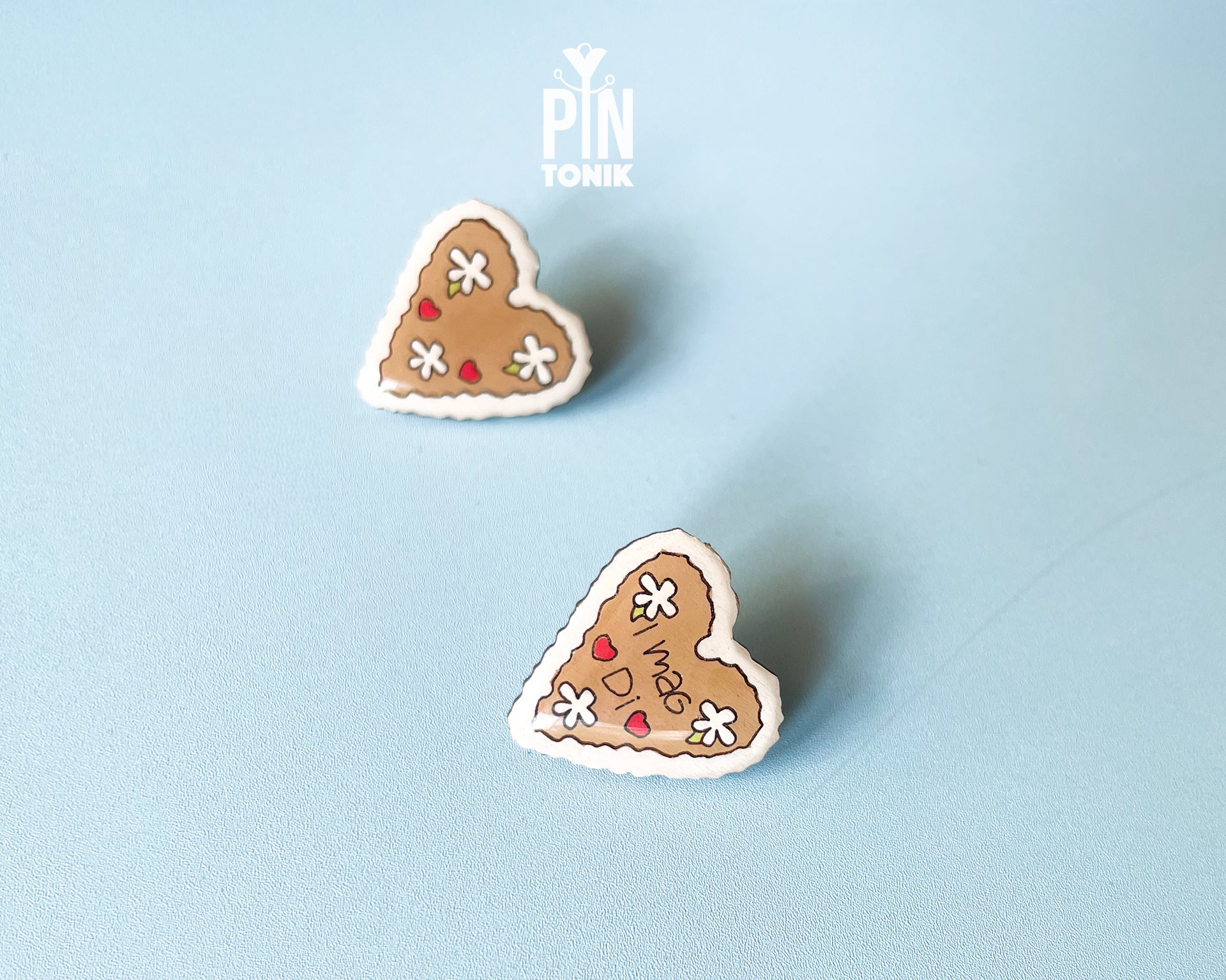 Hand-painted wooden gingerbread heart pins with white "icing" edges and floral details. One version features the Tyrolean text "I mag Di" while the other is a simple floral heart.