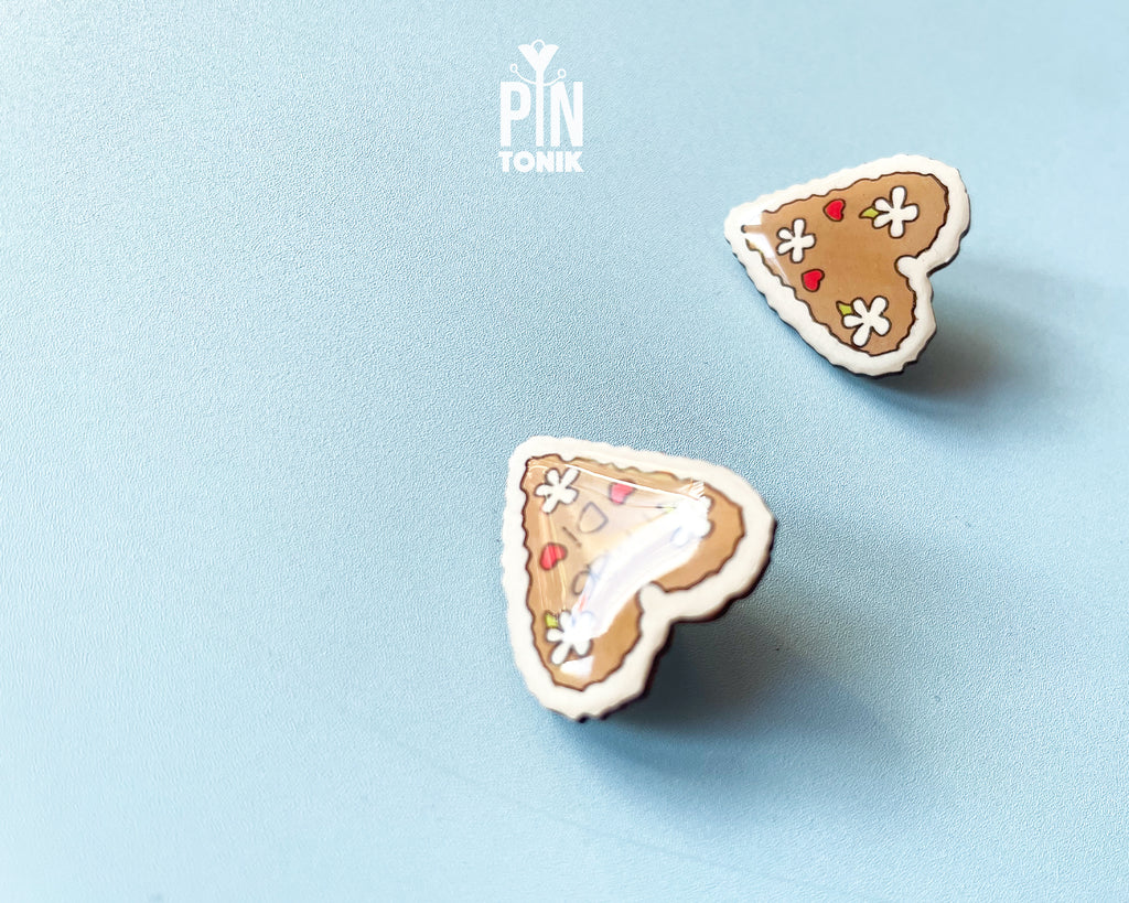 Hand-painted wooden gingerbread heart pins with white "icing" edges and floral details. One version features the Tyrolean text "I mag Di" while the other is a simple floral heart.