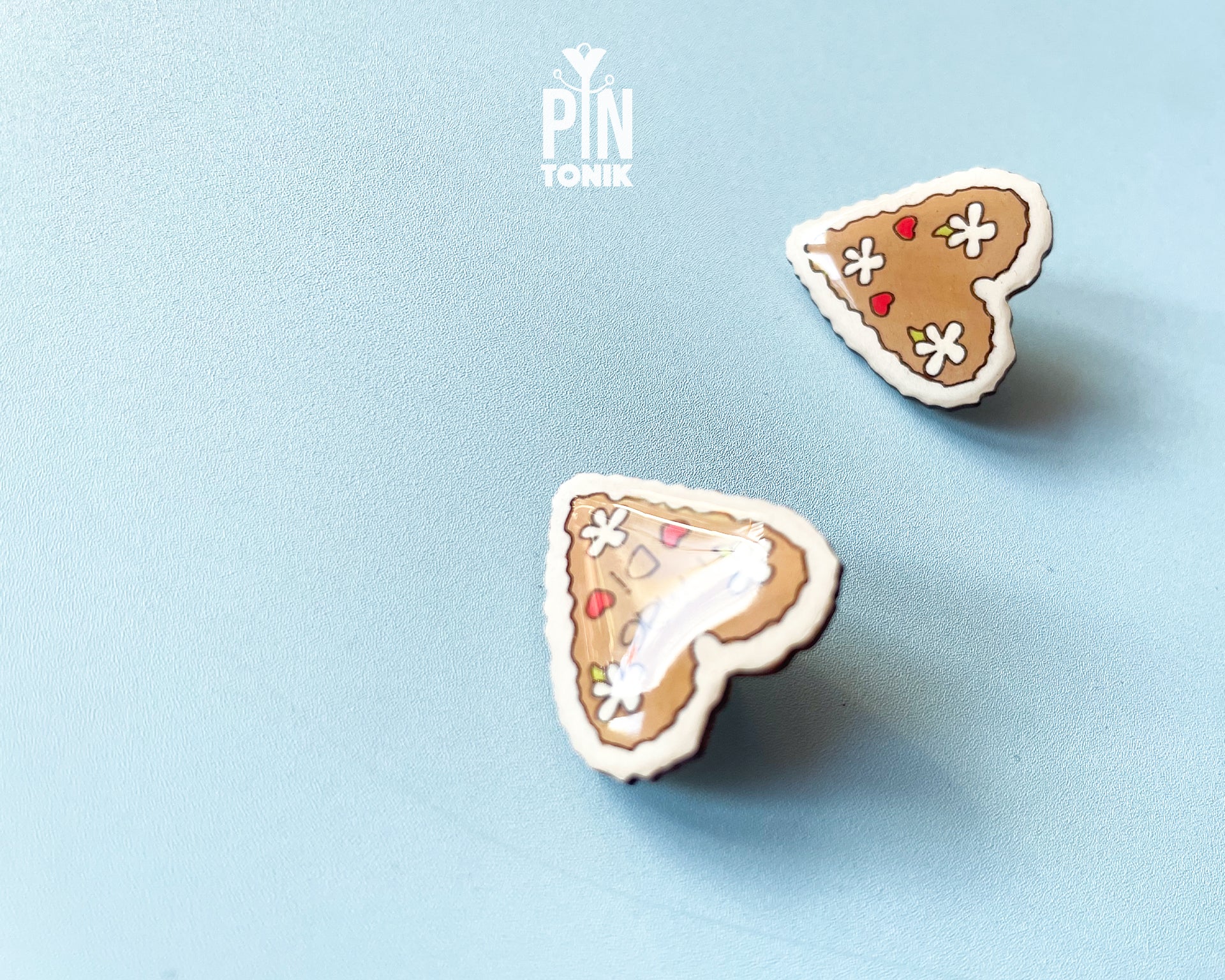 Hand-painted wooden gingerbread heart pins with white "icing" edges and floral details. One version features the Tyrolean text "I mag Di" while the other is a simple floral heart.