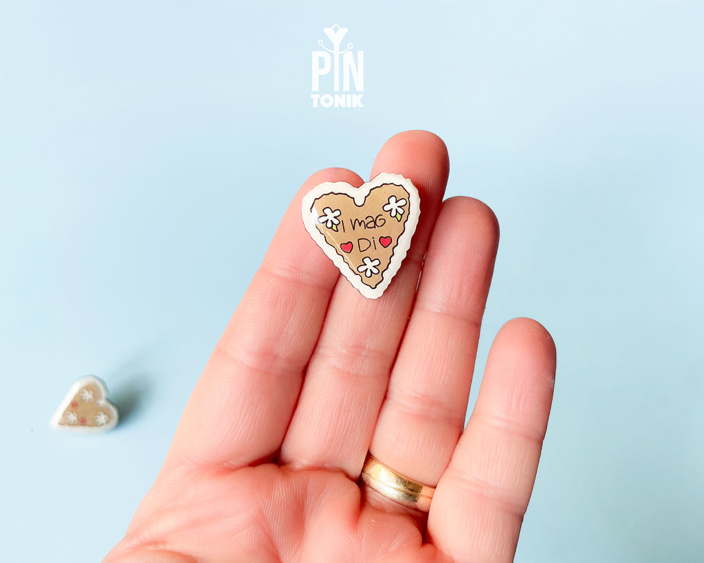 Hand-painted wooden gingerbread heart pins with white "icing" edges and floral details. One version features the Tyrolean text "I mag Di" while the other is a simple floral heart.