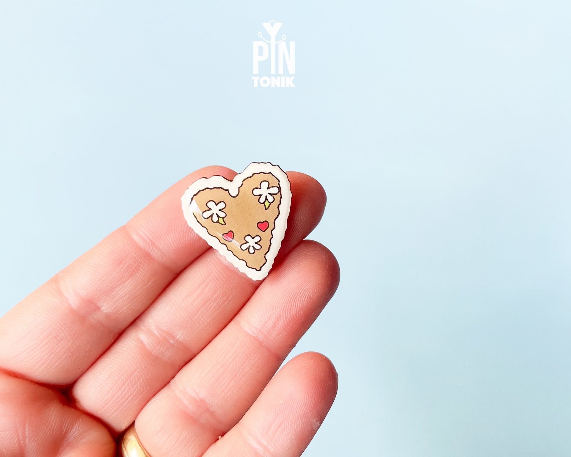 Hand-painted wooden gingerbread heart pins with white "icing" edges and floral details. One version features the Tyrolean text "I mag Di" while the other is a simple floral heart.