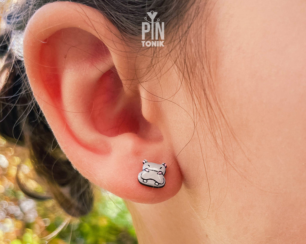 A close-up of a person's ear wearing a small white and black cat-shaped stud earring. The earring has a silver-toned outline with white enamel and black details.