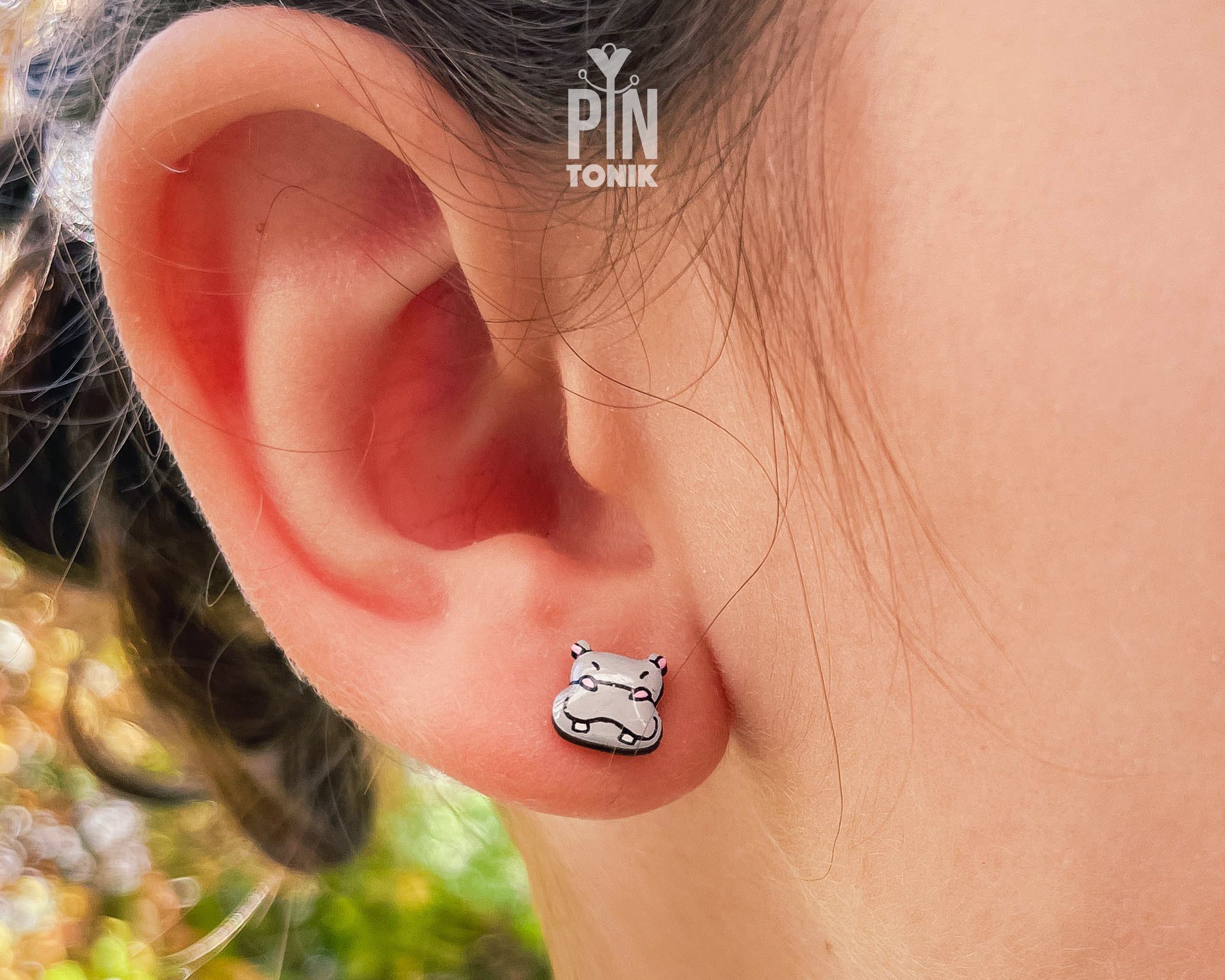 A close-up of a person's ear wearing a small white and black cat-shaped stud earring. The earring has a silver-toned outline with white enamel and black details.
